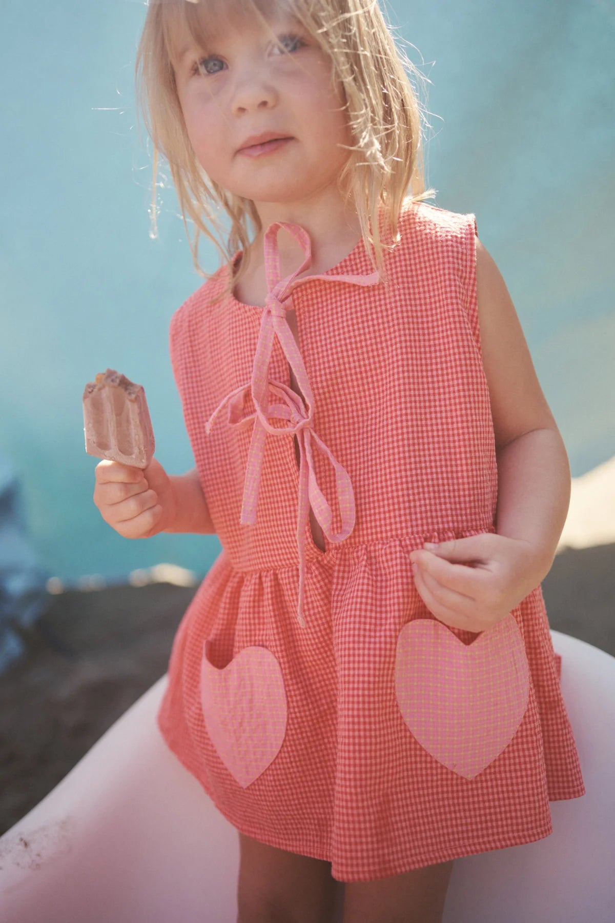 A young blonde girl stands outside in a Polly Pocket Blouse Cherry Gingham by LITTLE THE LABEL, featuring heart-shaped pockets. She holds a chocolate ice cream bar with a soft blue sky behind her, hinting at a sunny day.