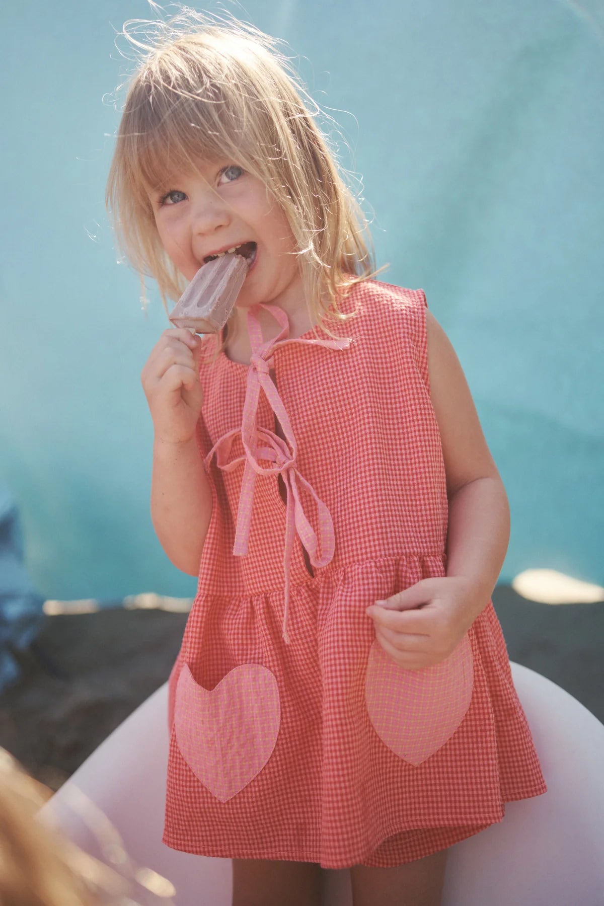 A young blonde child is happily enjoying a popsicle, dressed in the Polly Pocket Blouse Cherry Gingham by LITTLE THE LABEL, standing against a light blue background.