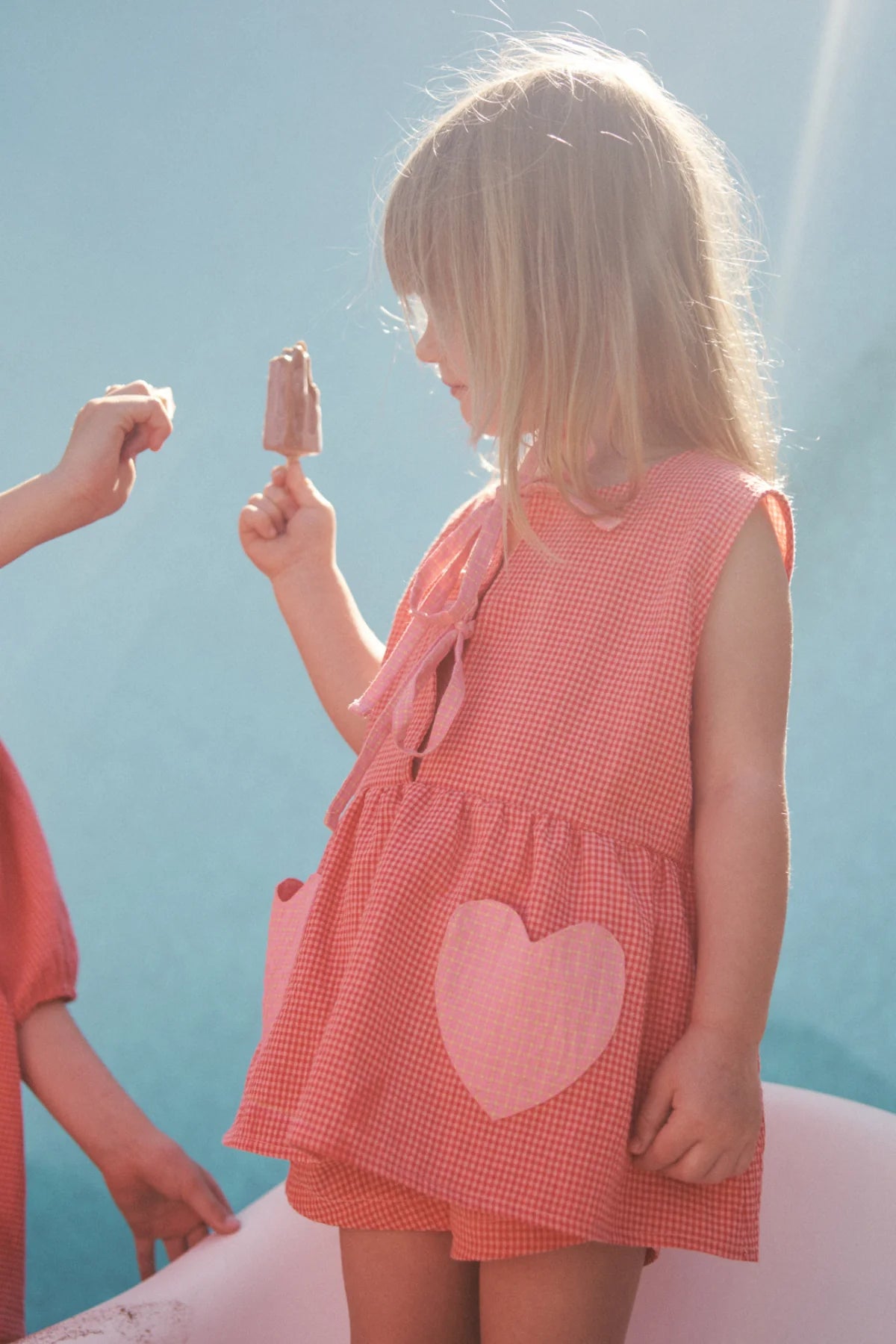 A young girl in a heart-patterned pink dress holds an ice cream bar in the sunlight against a blue backdrop, while another person reaches out from the left wearing LITTLE THE LABEL's Polly Pocket Blouse in cherry gingham, perfectly catching the light.