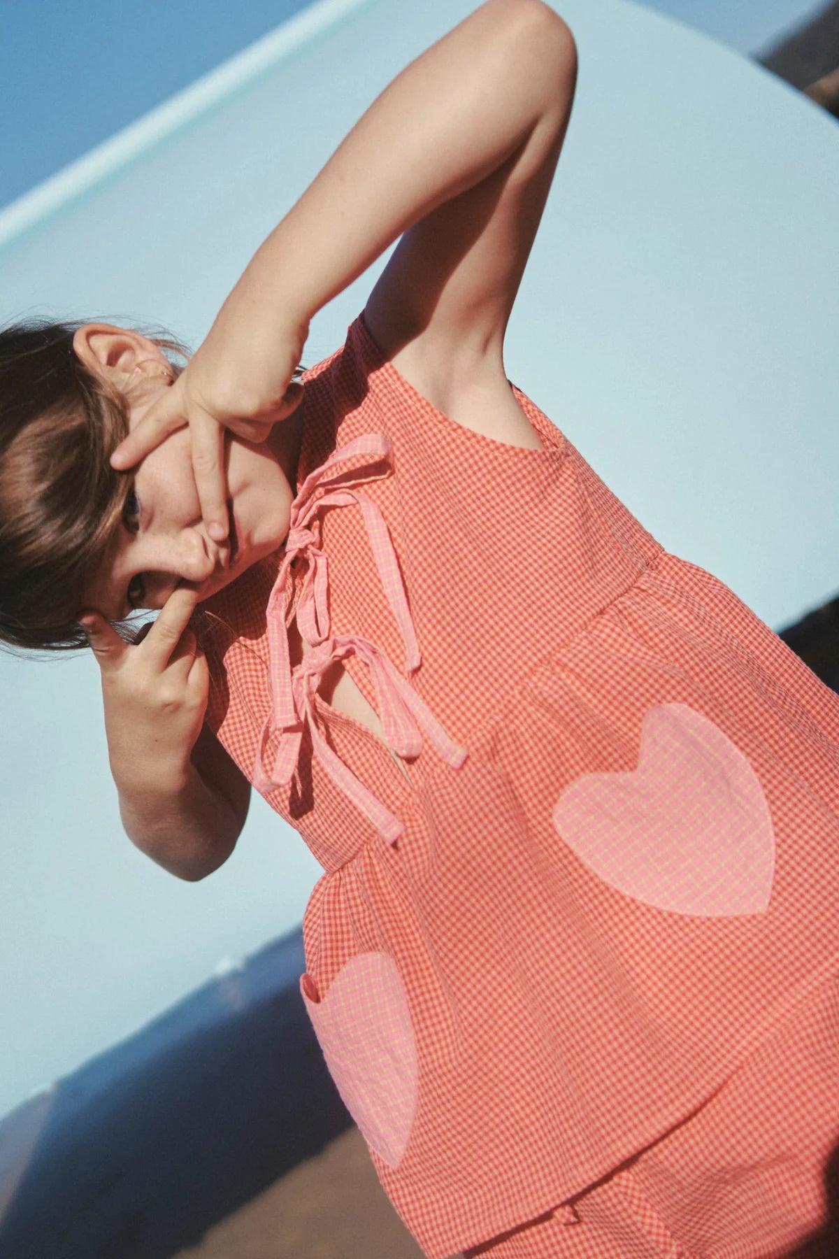 A young girl playfully poses in the Polly Pocket Blouse Cherry Gingham by LITTLE THE LABEL, framing her face with her hands against a clear blue sky, exuding a joyful and carefree vibe.