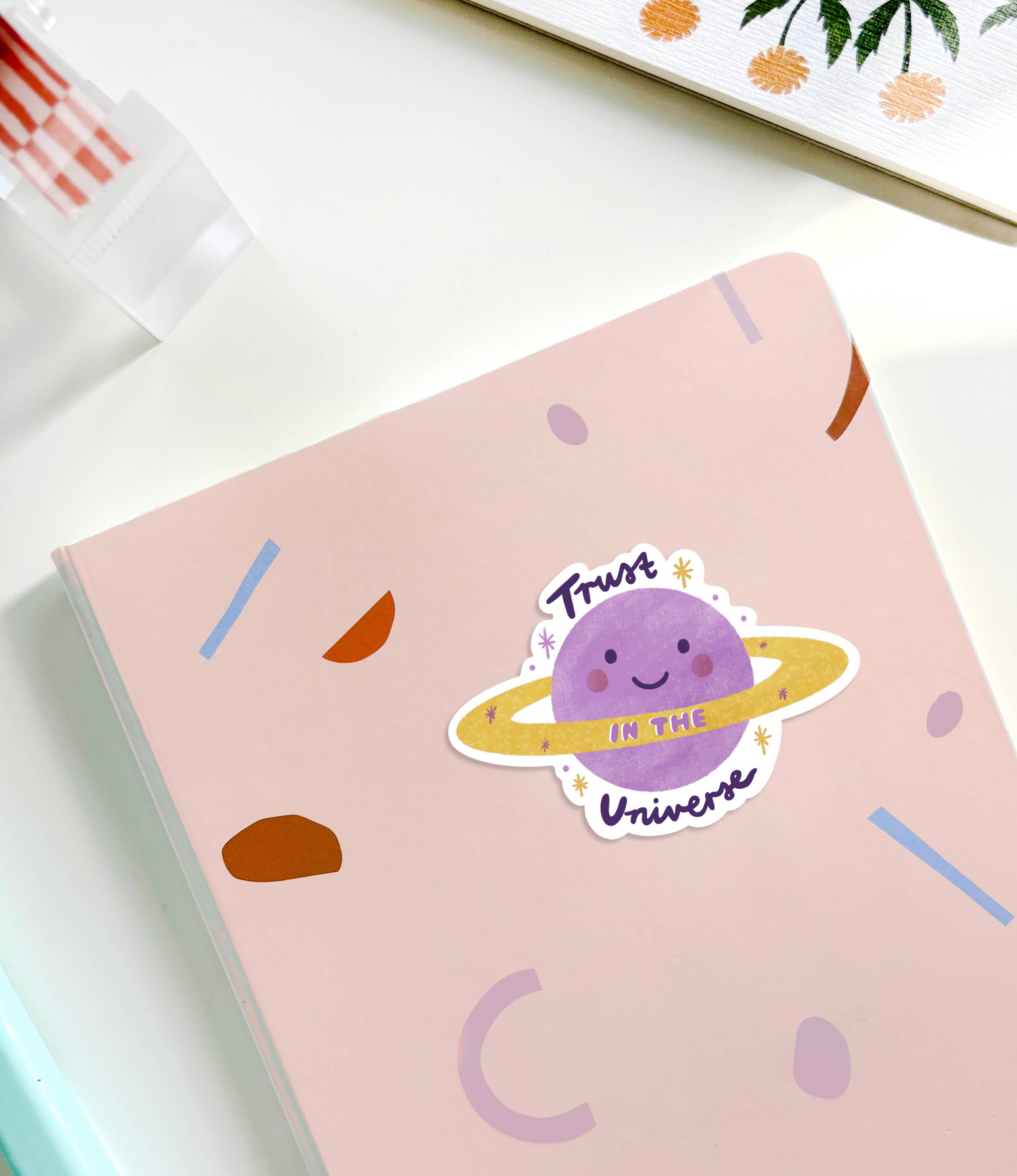 A pink notebook adorned with various geometric shapes on the cover sits on a white surface. At its center, a colorful Trust in the Universe Sticker from LAUREN SISSONS STUDIO showcases a smiling purple planet with a ring and uplifting text.