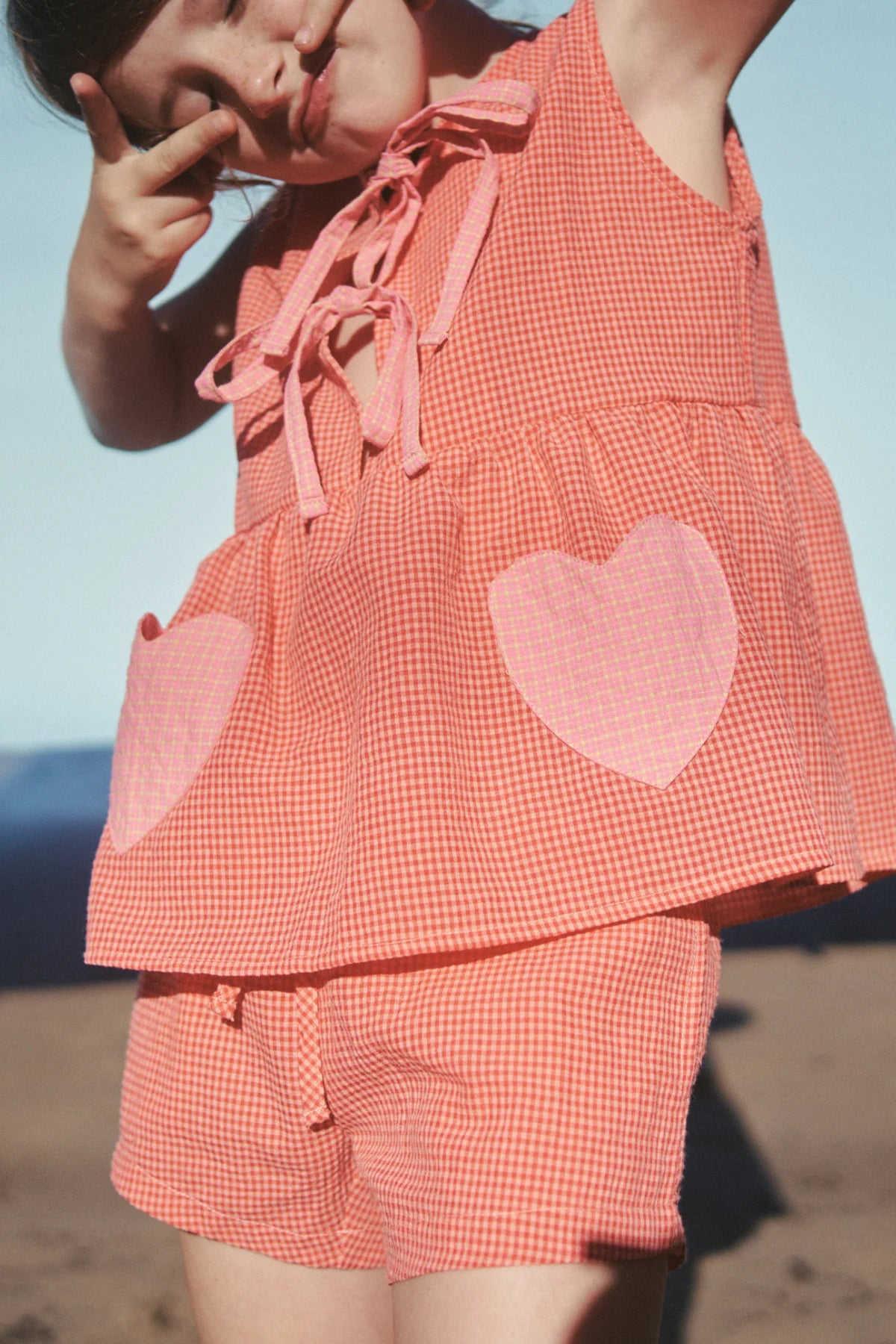 A child playfully poses against a clear blue sky in the Polly Pocket Blouse Cherry Gingham by LITTLE THE LABEL, featuring heart-shaped pockets.