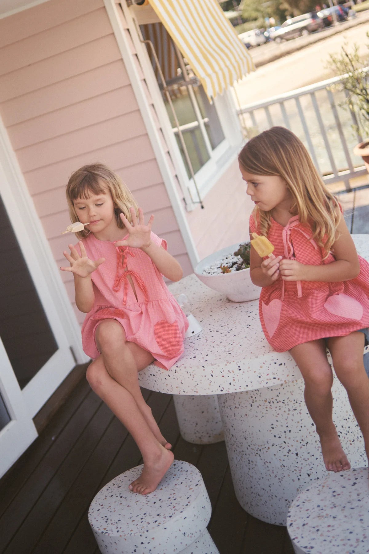 Two young girls in pink outfits sit outside on a terrazzo table and stools under a yellow-striped awning. One girl, wearing the Polly Pocket Blouse Cherry Gingham by LITTLE THE LABEL, licks an ice cream cone while the other gestures with her free hand, holding an ice cream bar.