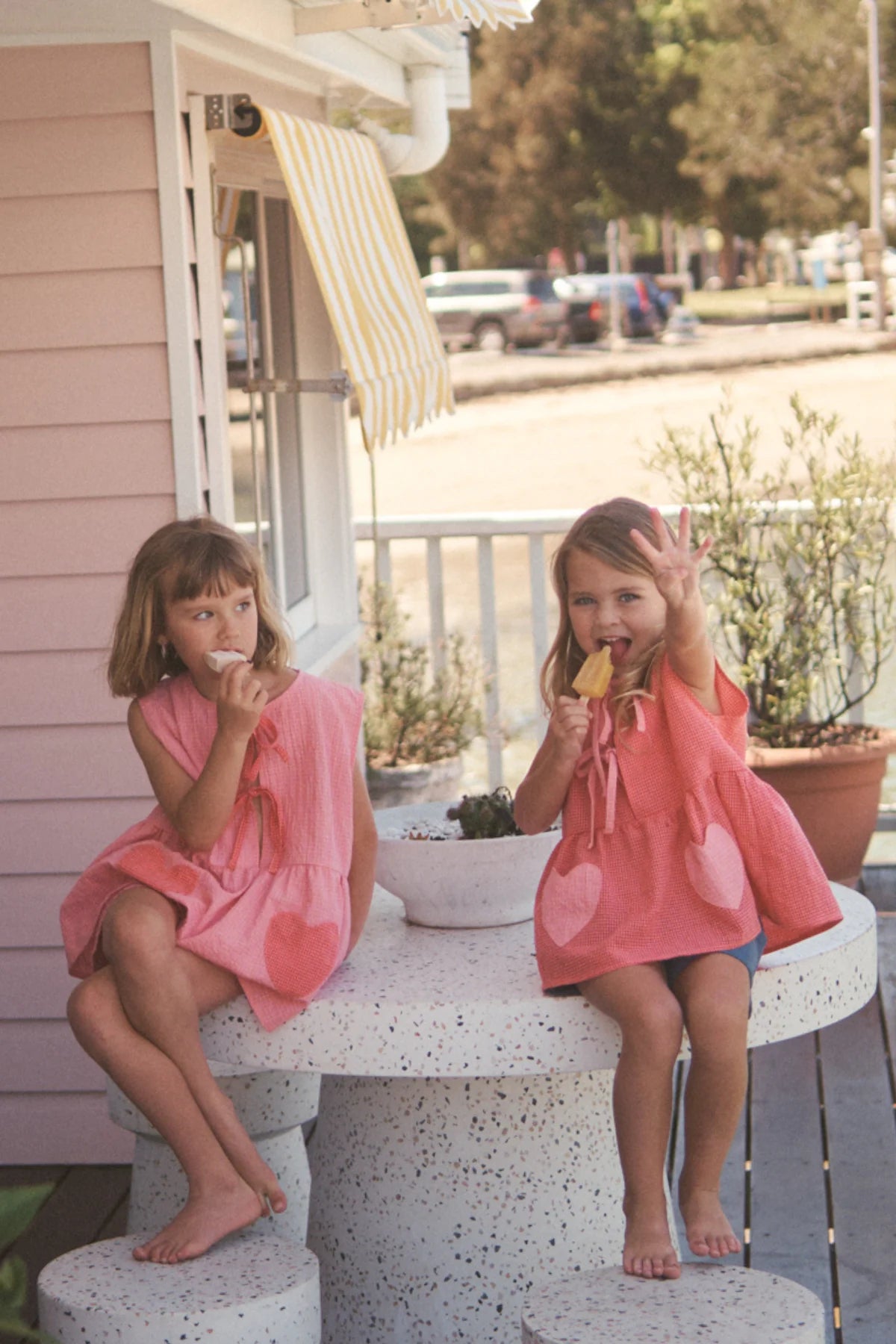 Two young girls in matching pink dresses sit on a stone table outdoors. One eats a snack, while the other waves with a smile. A small pink house with a yellow-striped awning and potted plants are in the background, evoking sunny days in LITTLE THE LABEL's Polly Pocket Blouse Cherry Gingham.