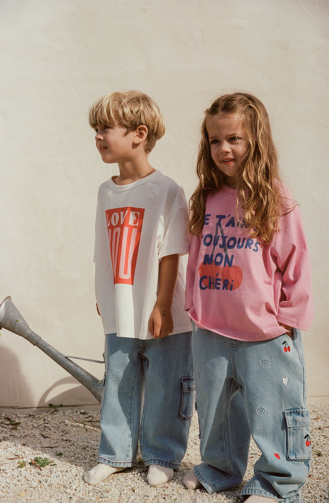 Two young children stand together outdoors against a plain wall. The boy is dressed in a white T-shirt with a red "LOVE YOU" graphic paired with blue cotton denim jeans, while the girl wears a pink long-sleeve shirt featuring French text and wide-leg Louie Denim Jeans Mon Cheri from THE WHOLESOME STORE. Both are barefoot and appear happy.