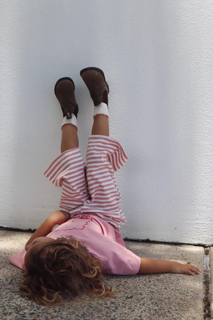 A child outdoors with brown boots and floaty pants in candy cane pink stripes by BOBBY G BABY WEAR, lies on their back with legs against a white wall, paired with a pink shirt. The ground is textured.