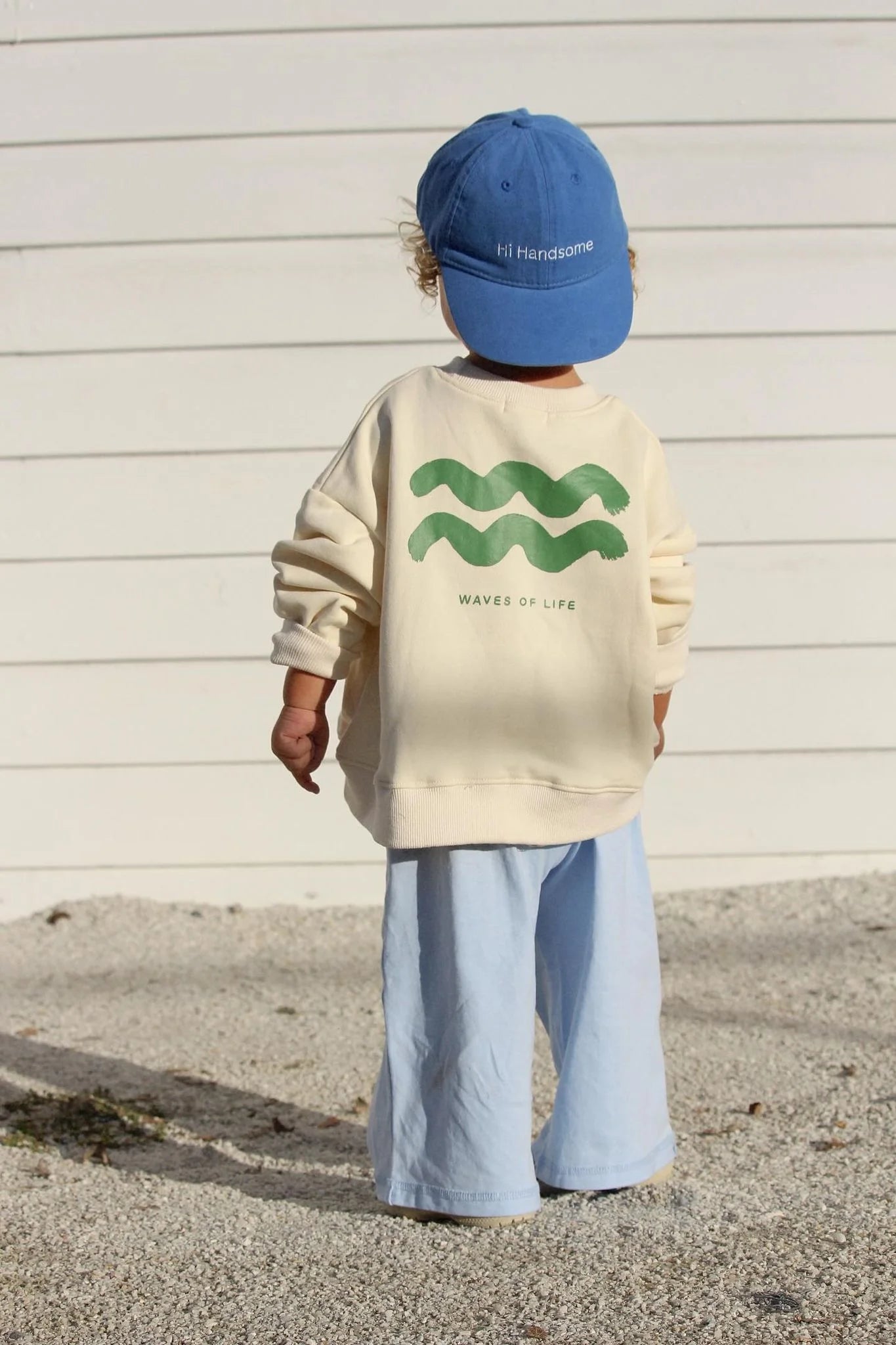 Young boy wearing the wave of life pullover in creme and a blue cap and pants.