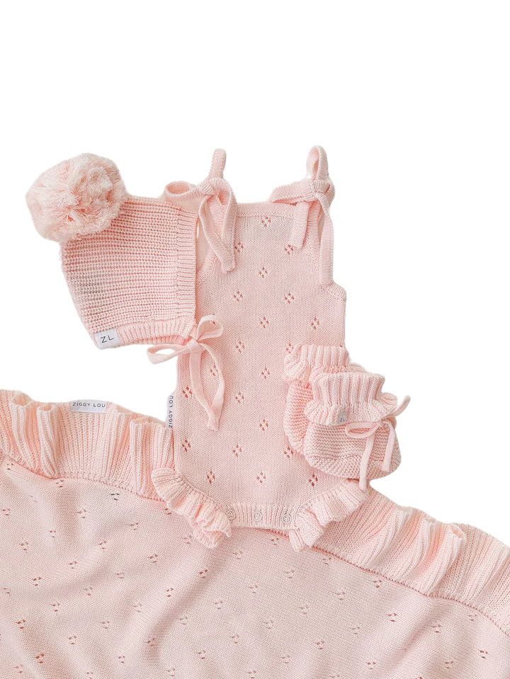 Presenting the Frill Bodysuit Primrose from ZIGGY LOU, a delightful coordinated baby outfit set in light pink. This charming ensemble includes a knitted frill bodysuit with bow details, a matching bonnet with a pom-pom, and ruffled booties. Part of the Primrose Signature Collection, it is crafted from 100% cotton and adorned with delicate eyelet patterns and ruffled edges.