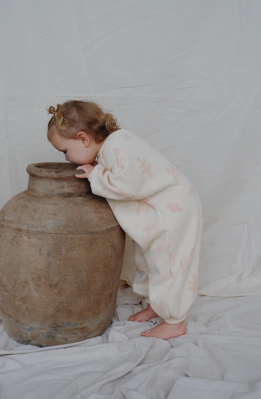 A little girl wearing the Myla Jane fleecey romper in peach while looking into a ceramic pot.