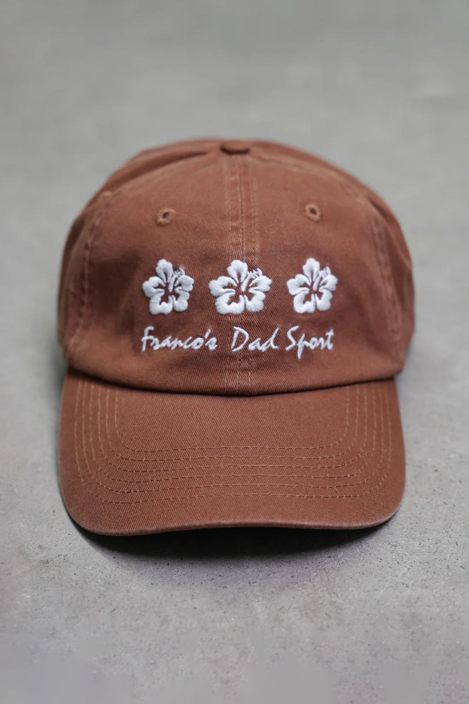 A "PRE-ORDER Hibiscus Cap" in brown, featuring three intricately embroidered white flowers and the words "FRANCO'S DAD Sport" on the front, is showcased on a gray surface.