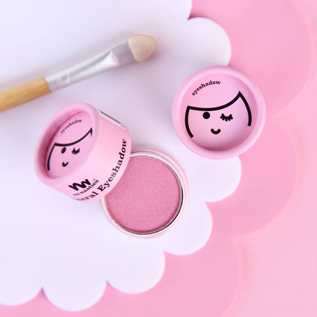 An open container of Kids Eco Natural Pressed Eyeshadow & Blush by no nasties kids, featuring a small round hypoallergenic pink eyeshadow with a cartoon face design on its lid, reveals the product inside. A matching applicator brush with a bamboo handle rests next to it. Both cruelty-free items are displayed on a white and pink scalloped mat, set against a pink background.