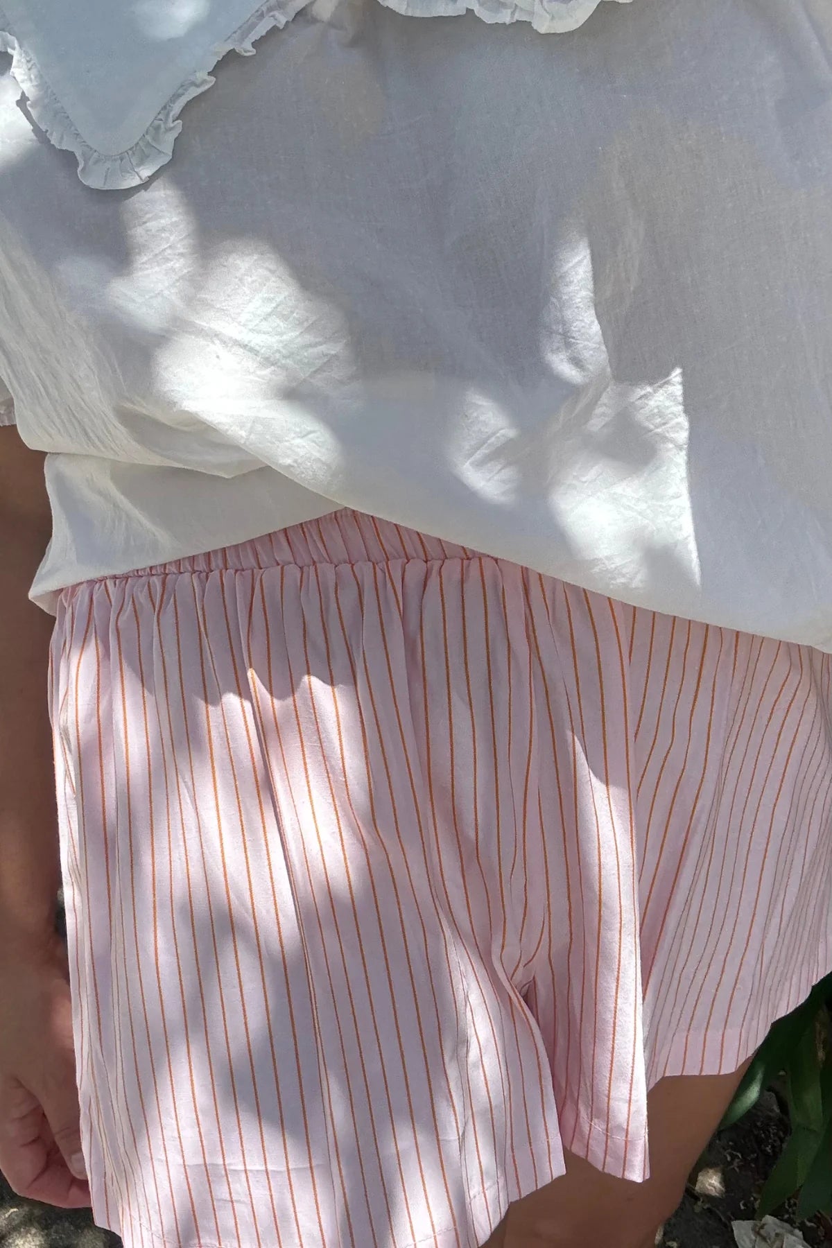 Wearing a white ruffled blouse and LITTLE THE LABEL's Mama Everyday Shorts Sunset Stripe, sunlight casts dappled shadows that add an organic pattern to the fabric, capturing the effortless charm of Mama Styles.