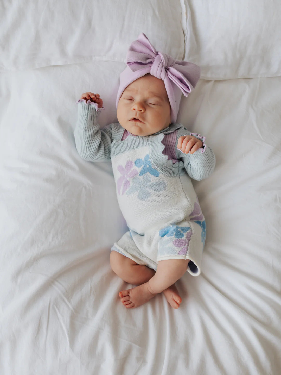 A sleeping baby lies on a white bed, dressed in a light blue ZIGGY LOU Playsuit Alaska with a floral pattern and a matching large lavender bow headband. The baby’s hands are gently positioned, and feet are partially visible. The background is simple and neutral, emphasizing the baby.