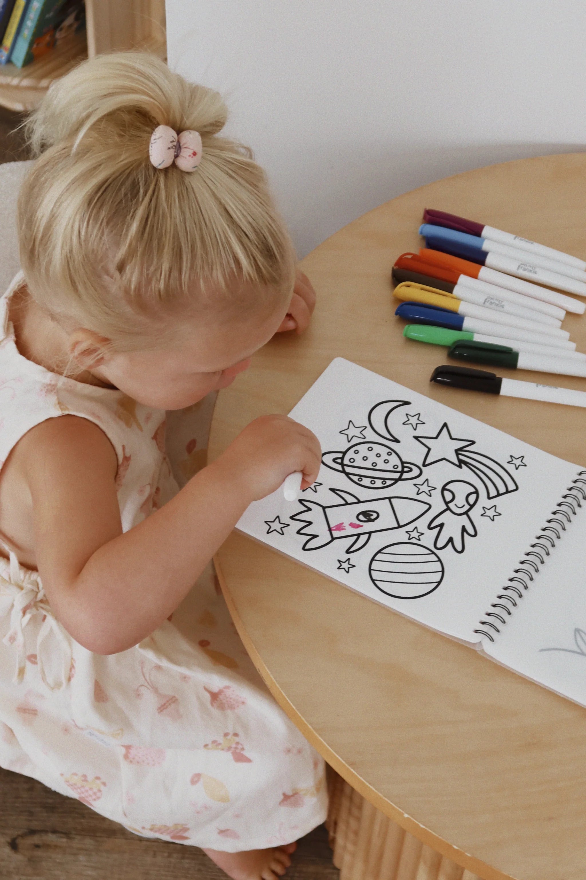 A blonde girl in a ponytail, wearing a floral dress, colors the Oh My Frankie ~ Oh My Scribbles Reusable Colouring Book with non-toxic markers at a round wooden table. She focuses on drawings of planets, stars, and a rocket.