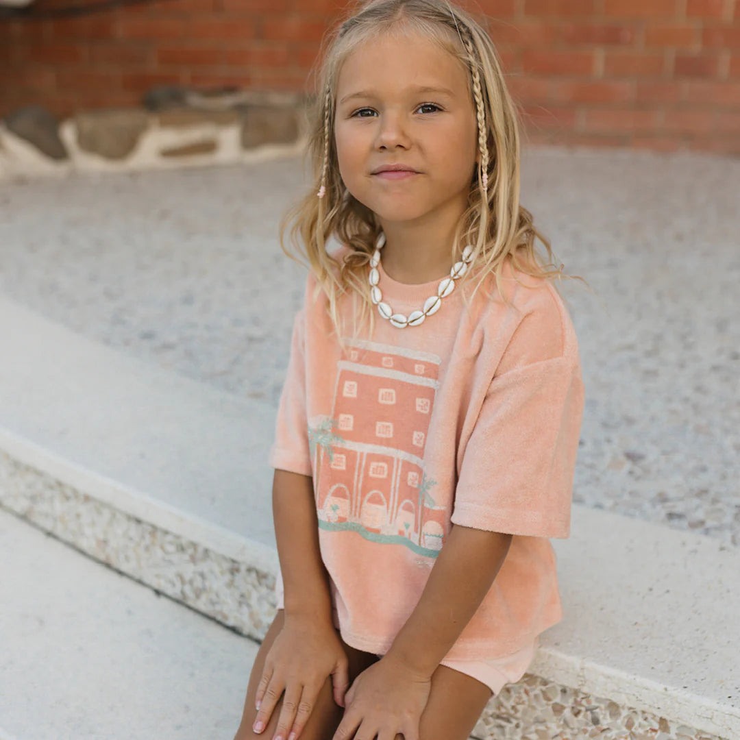 A young child with light hair, wearing a peach-colored Room Service Mid Sleeve Tee Terry Towel Spanish Villa by GOLDEN CHILDREN, adorned with a shell necklace, sits outdoors on a textured surface. The background features a brick wall and partial stonework from the nostalgic Colony Hotel.