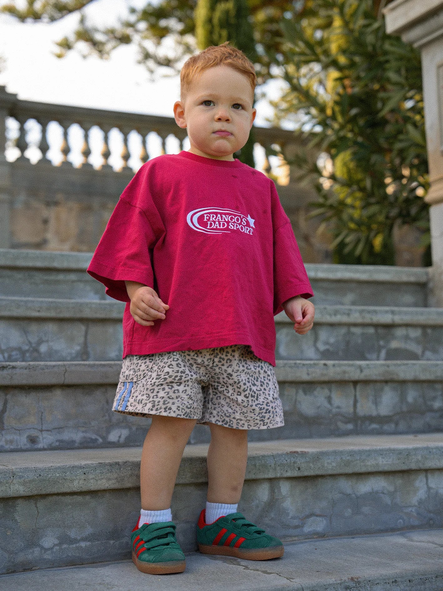 A young child stands on stone steps wearing an oversized red t-shirt, a pair of PRE-ORDER Leopard Cargo Shorts by FRANCO'S DAD, and green shoes with red stripes. Trees and a balustrade provide a lush backdrop.