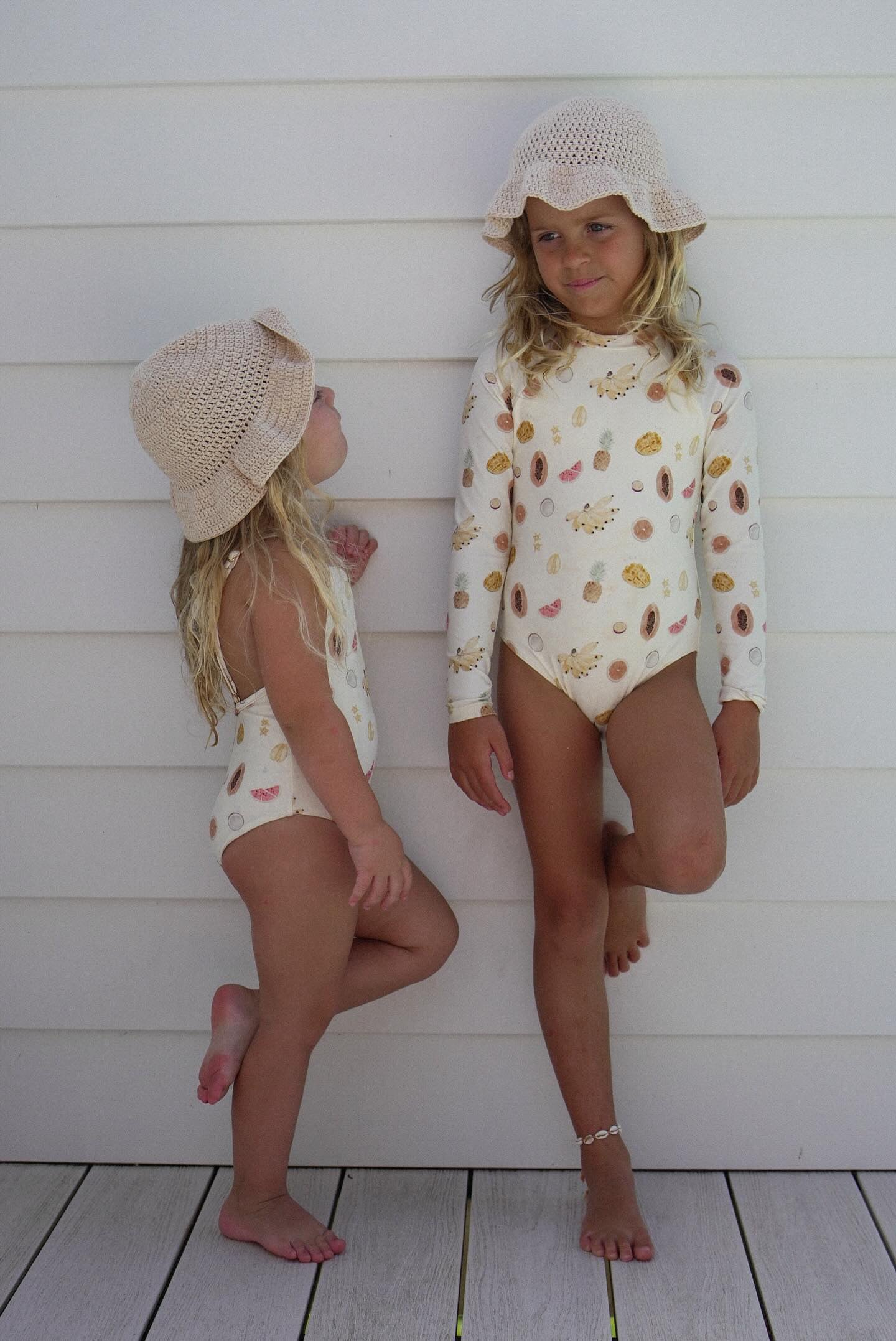 Two young girls stand against a white wooden wall, both donning the Tutti Frutti One Piece long-sleeve toddler swimsuits with adjustable straps from EN. THE LABEL and matching beige sun hats. The older girl rests one leg on the wall, glancing down, while the younger girl gazes up at her with one leg slightly bent.