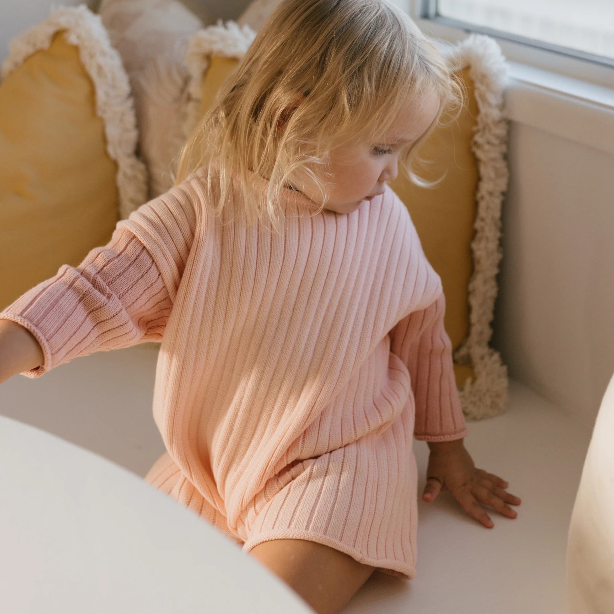 A young child with blonde hair is sitting on a white surface near a window, wearing the Golden Knit Romper Strawberry Sundae by GOLDEN CHILDREN, made from 100% cotton knit. Behind the child are two yellow pillows with fringed edges. The child looks down at their hand placed on the surface, showcasing their stylish transeasonal all-in-one piece.