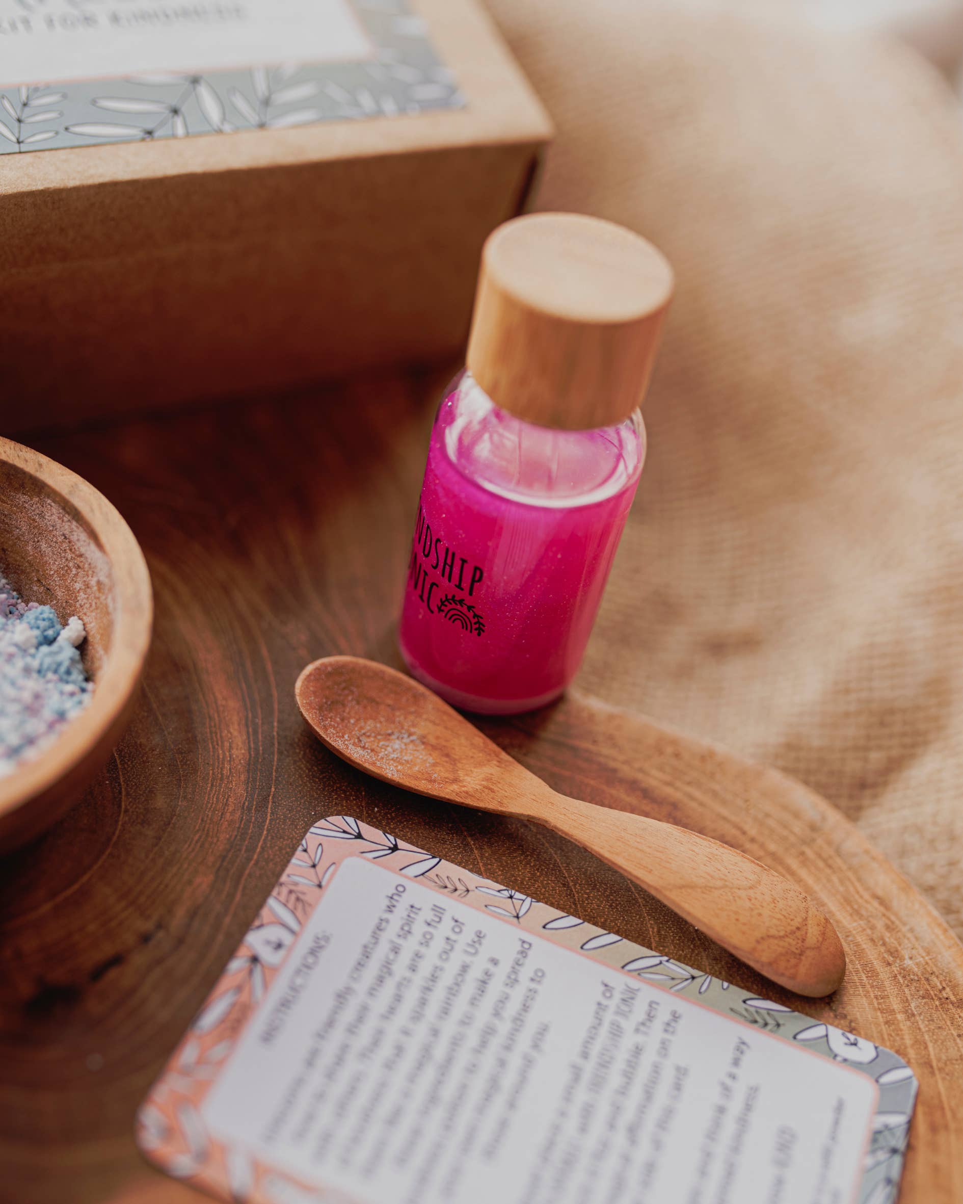 A small bottle of bright pink liquid with a wooden cap, a small wooden spoon, a bowl filled with colorful contents, and an artfully arranged card with text sit on a wooden surface. In the background, a partially visible cardboard box adorned with a leaf design hints at the MINI Rainbow Sparkles Potion Kit (A potion for kindness) by THE LITTLE POTION CO.