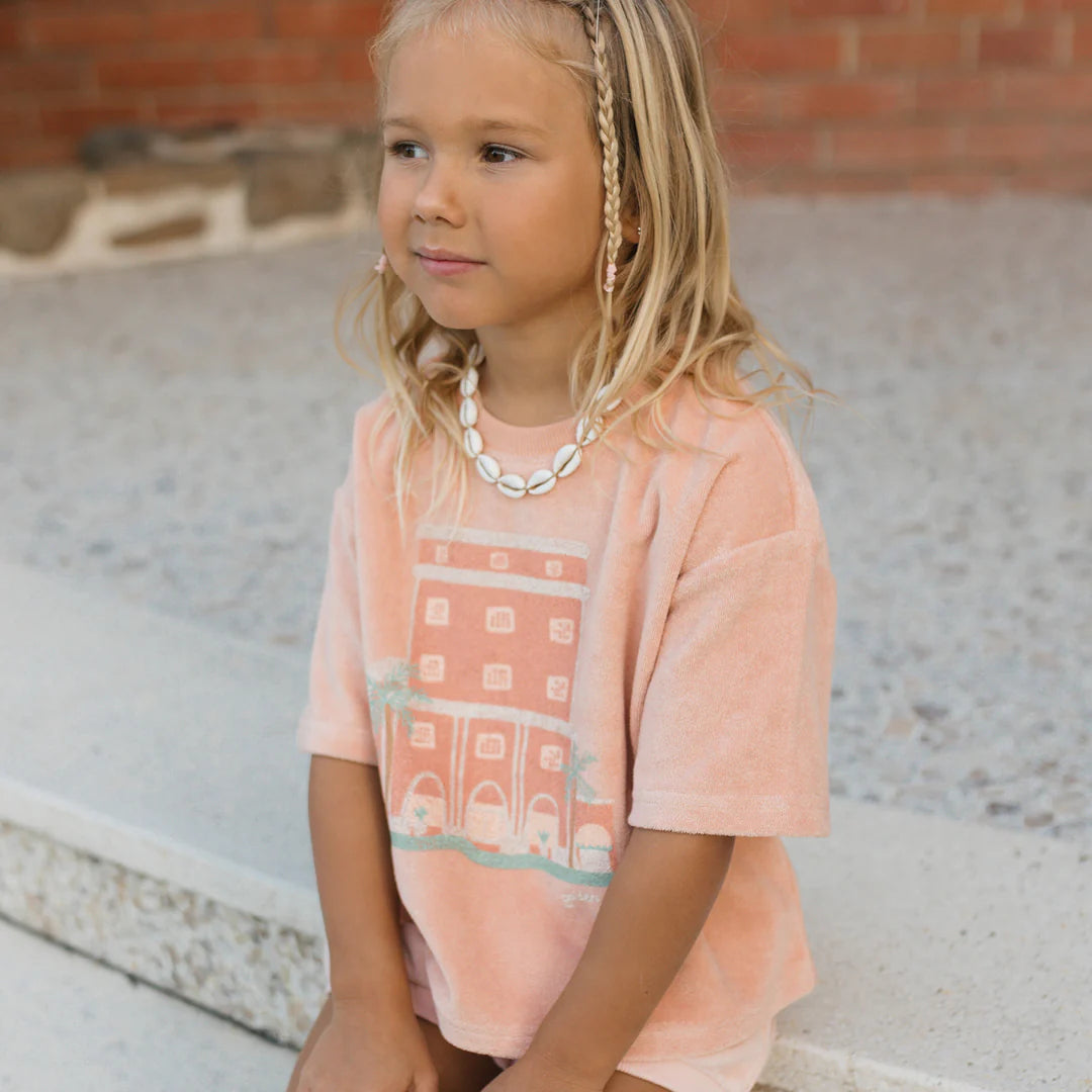 A young girl with blonde hair sits on concrete steps, dressed in a peach-colored Room Service Mid Sleeve Tee Terry Towel Spanish Villa by GOLDEN CHILDREN, featuring a building design. Her light shorts complete the ensemble, and she accessorizes with a beaded necklace. Small braids embellish her hair as she looks to the left, with a brick wall providing the backdrop.