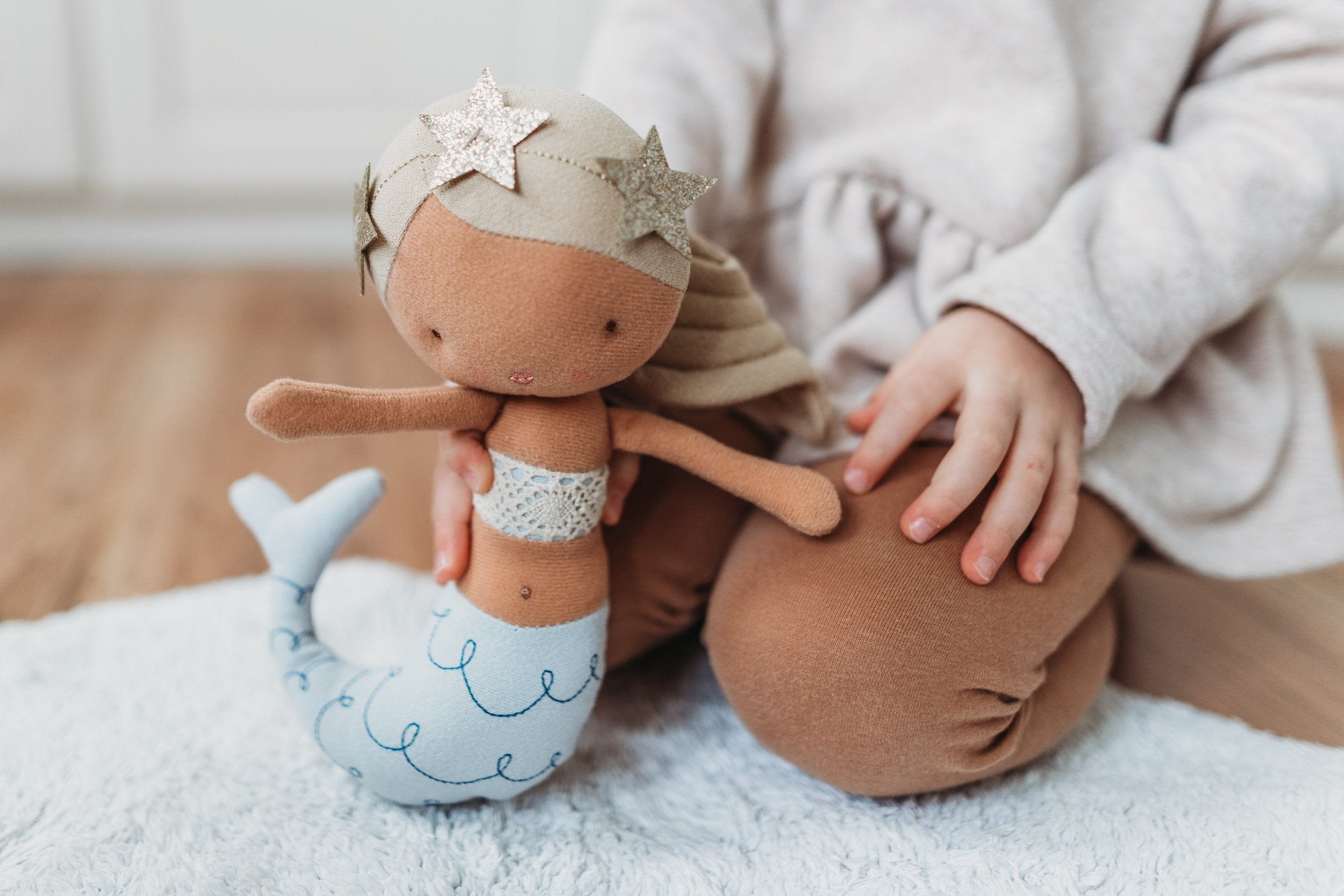 A child in a beige sweater and brown leggings sits on a light blue rug, gently holding PICCA LOULOU's Mermaid Pearl 22cm, a fabric mermaid doll with blond yarn hair, a sequined top, a light blue tail adorned with swirly patterns, and sparkly star decorations.