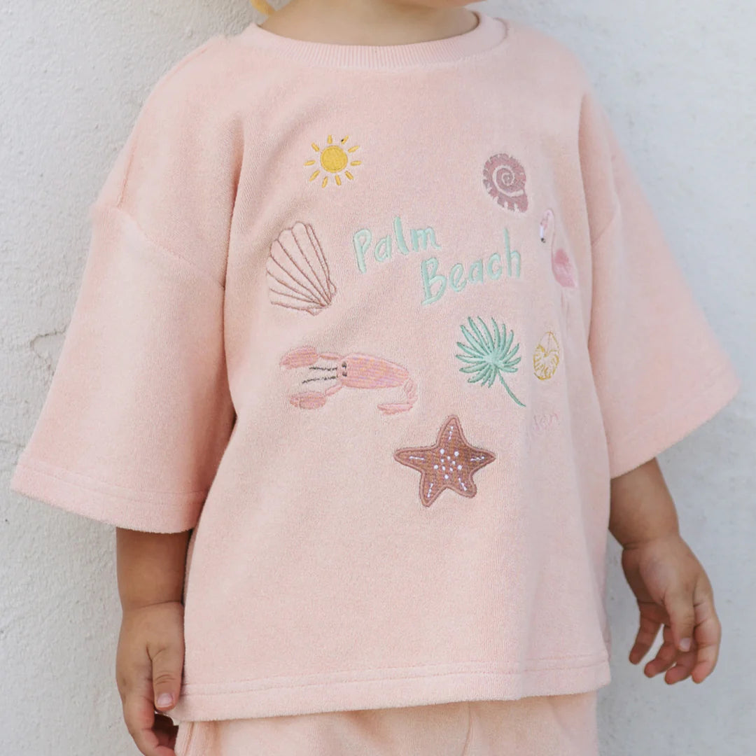 A child is dressed in the comfortable Palm Beach Mid Sleeve Tee Terry Towel Flamingo Pink by GOLDEN CHILDREN, showcasing detailed embroidery of a sun, seashell, lobster, palm leaf, and starfish. Made from soft terry towel cotton for optimal comfort, this shirt features a delightful light pink hue.