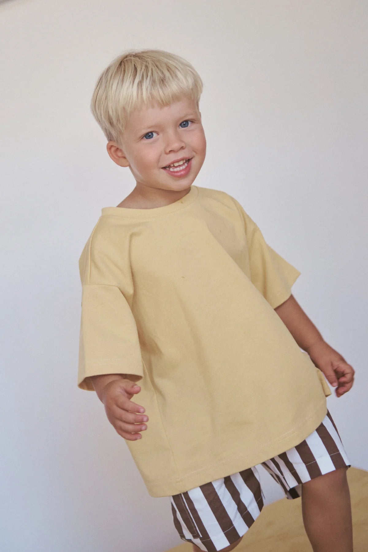 A young child with blonde hair smiles while wearing an oversized Little Lou Tee Pomello from LITTLE THE LABEL, paired with brown-and-white striped shorts, standing against a plain background.