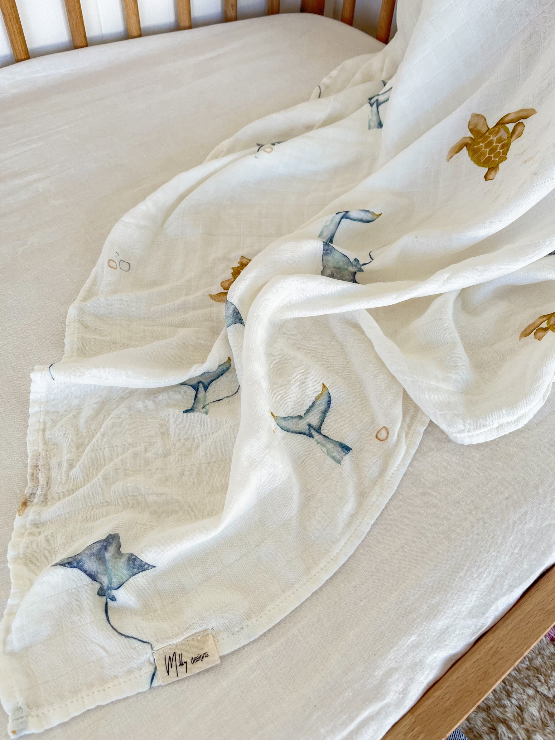 A white baby crib with a soft Milky Designs ~ Sea Creatures Wrap by MILKY DESIGNS draped over the mattress. This breathable blanket features blue manta rays and green turtles, with a small logo tag on its edge.