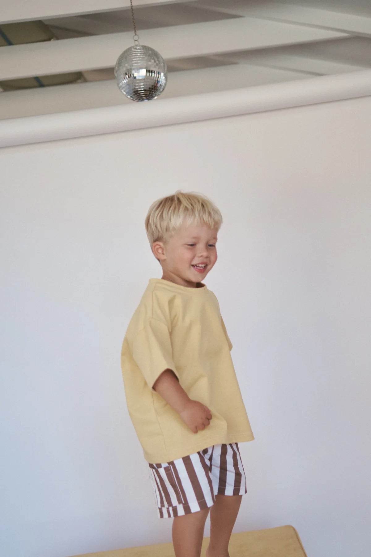 A young child with blond hair joyfully stands on a wooden platform, wearing an oversized Little Lou Tee Pomello by LITTLE THE LABEL and striped shorts. The room's disco ball and plain white background enhance the bright, cheerful vibe.