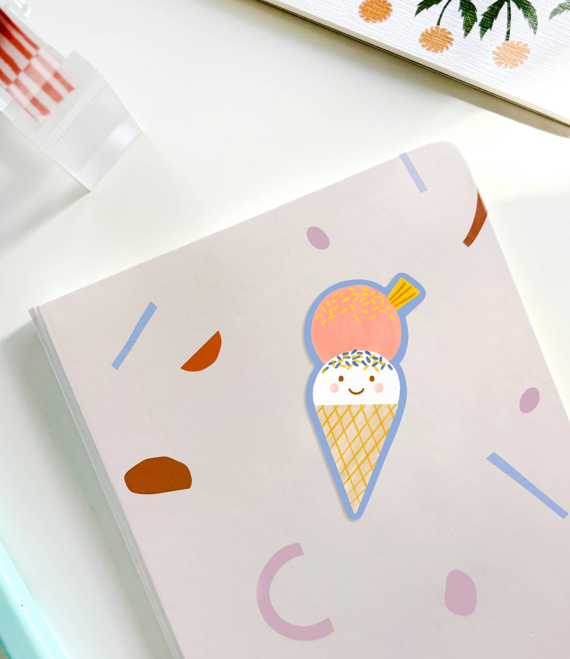 A pastel-colored notebook with a cute ice cream cone illustration on the cover. The ice cream, part of the Ice Cream Sticker collection from LAUREN SISSONS STUDIO, features a smiling face with two scoops, one pink and one white with sprinkles. The background showcases abstract shapes in various pastel colors, all protected by UV-coated matte vinyl for durability.