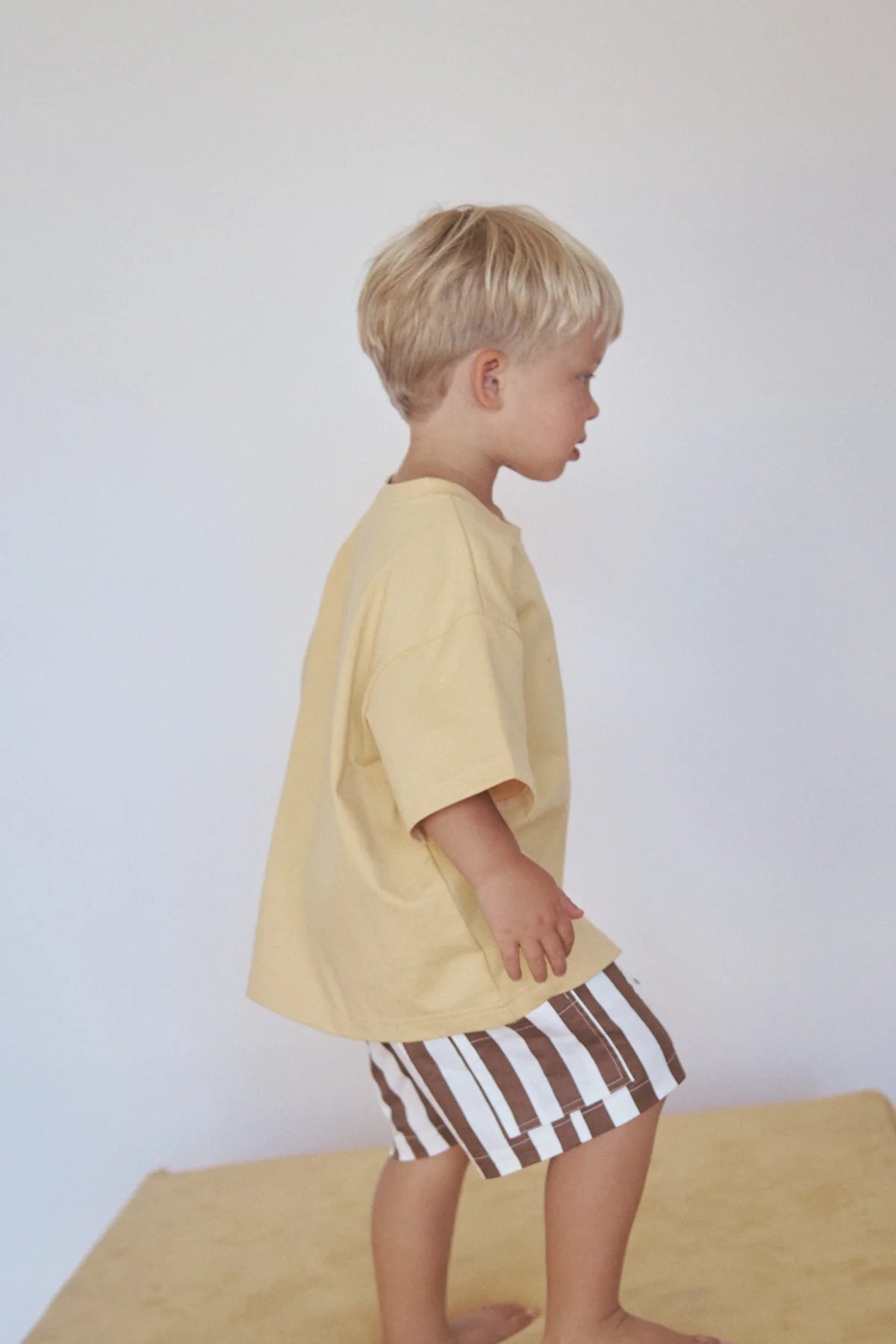 A young child with blonde hair stands on a light surface against a plain white backdrop, wearing an oversized fit Little Lou Tee Pomello by LITTLE THE LABEL in 100% cotton and brown-and-white striped shorts, gazing thoughtfully to the side.