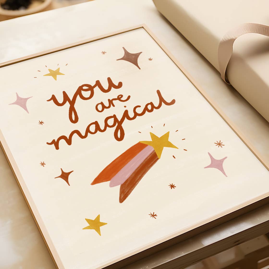 An art print set on a beige background, hand-illustrated type reads "You are Magical" surrounded by tiny sparkles and a shooting star by Laura Sissons Studio.