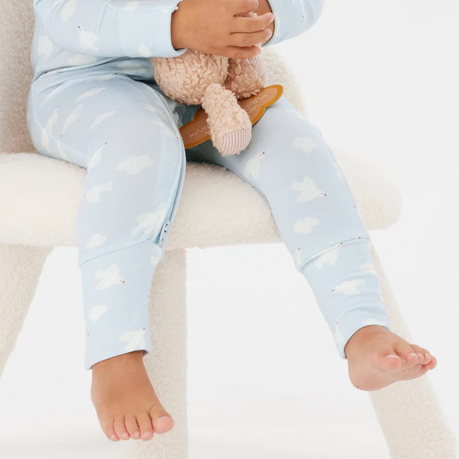 A child in the Day or Night Onesie Bird Song from KYND BABY, featuring a cozy light blue design with cloud patterns, sits on a white chair while holding a plush toy and rubber teething ring. The onesie, crafted from sustainable EcoCosy® Viscose fabric, reveals their lower legs and bare feet.