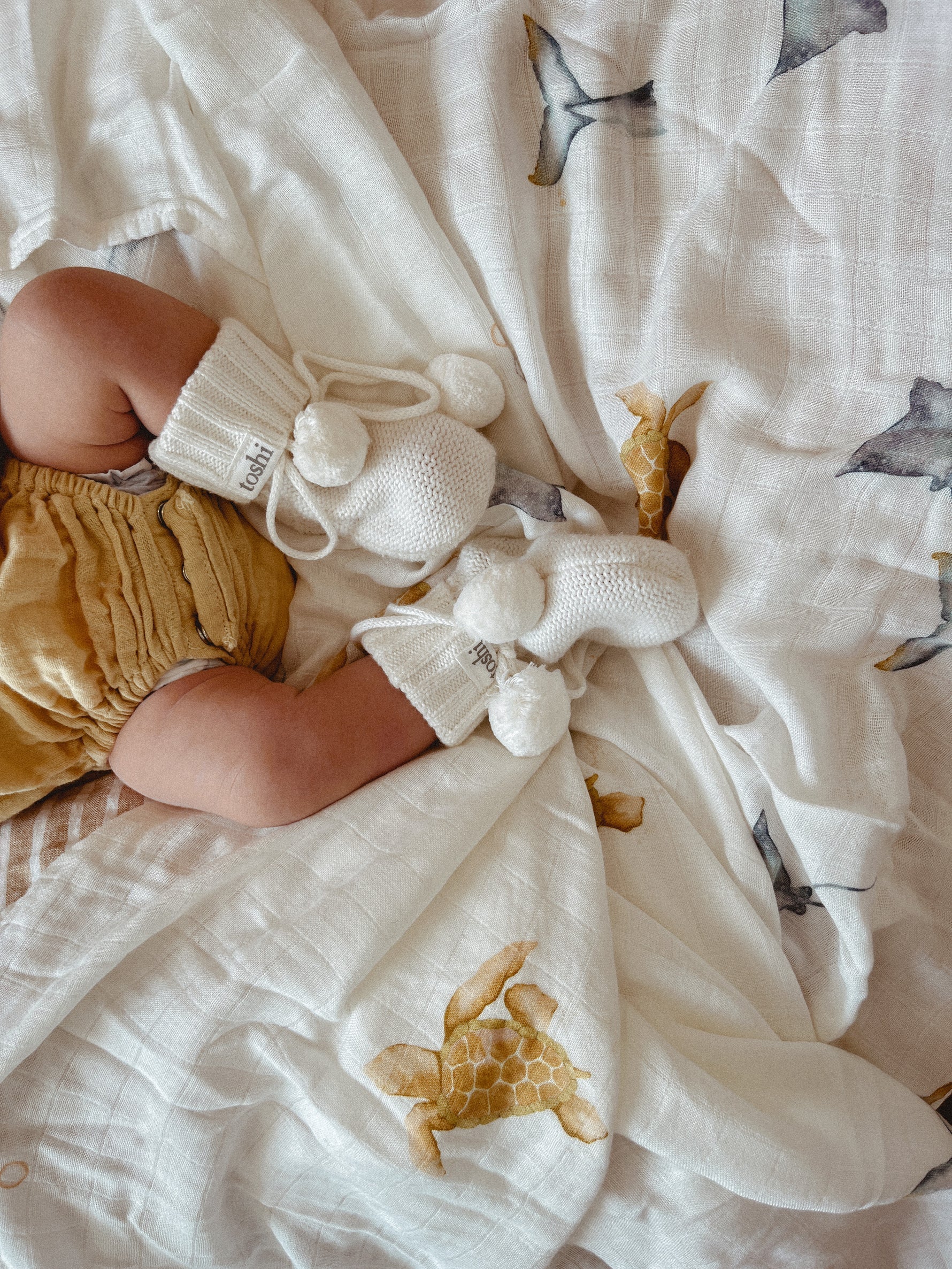 A baby rests on the Milky Designs ~ Sea Creatures Wrap by MILKY DESIGNS, swathed in an outfit of exceptional breathability. Cream-colored knit booties with pom-poms complete the cozy scene, enveloping the little one in warm tranquility.