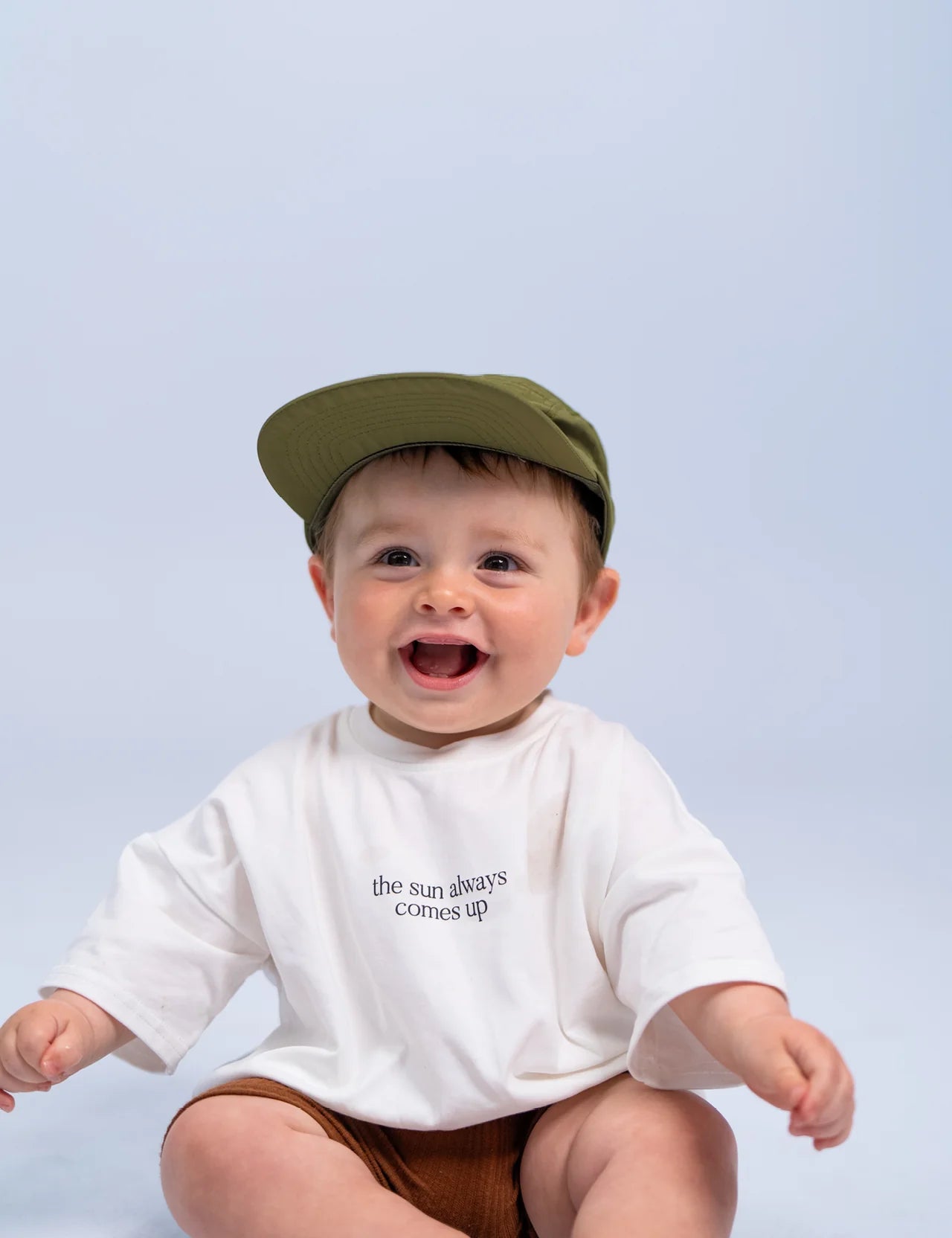 A baby, sitting and smiling, wears a SONNY LABEL Summer Club Cap in khaki with an adjustable velcro strap, paired with a white T-shirt featuring the phrase "the sun always comes up." The soft, light gray background perfectly captures the baby's joyful and content demeanor.