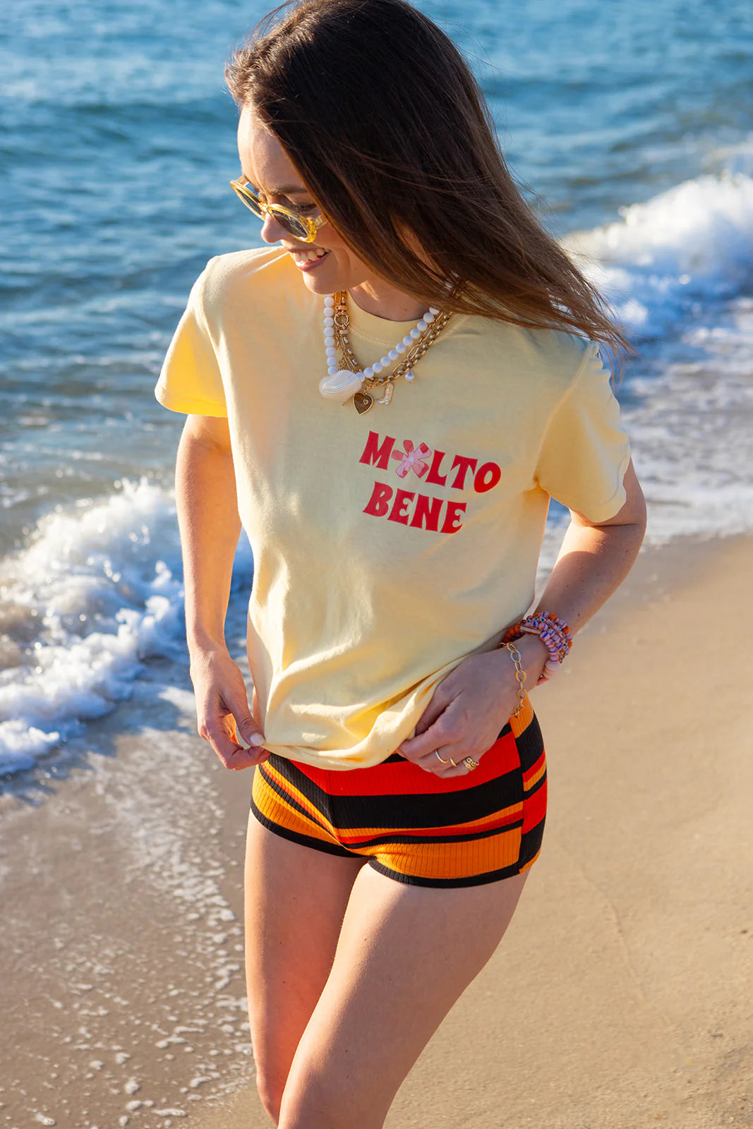 A woman in a 100% cotton light yellow "Molto Bene" crop tee by MAKU THE LABEL and striped shorts smiles as she walks along the sunny beach. Sunglasses shield her eyes while ocean waves gently touch her feet.