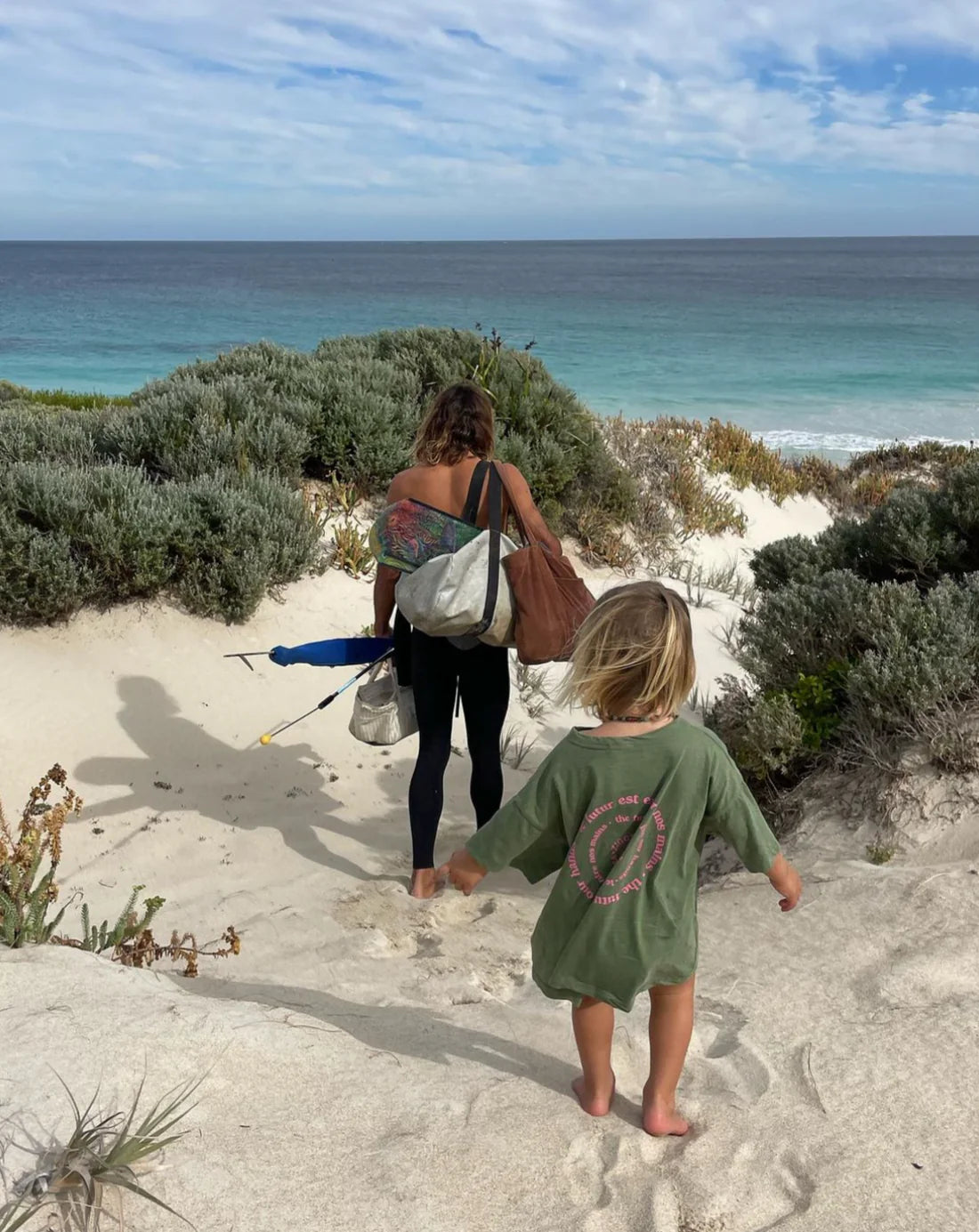 A woman and child walk down a sandy path to the beach with bags and a beach umbrella. The child wears ZIGGY ZAZA's "Ziggy Zaza ~ Love Your Mother Tee Lizard," showcasing its relaxed boxy fit, while the woman sports leggings. Ocean waves roll under a partly cloudy sky.