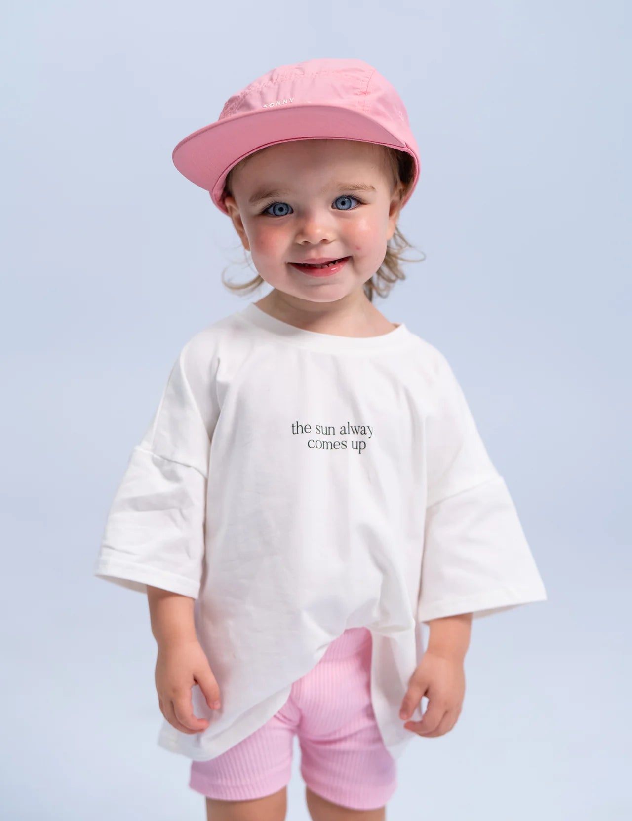 A young child wearing the Summer Club Cap: Flamingo by SONNY LABEL, a pink cap with an adjustable Velcro strap, along with a white t-shirt that says "the sun always comes up" and pink shorts, stands smiling against a light blue background.