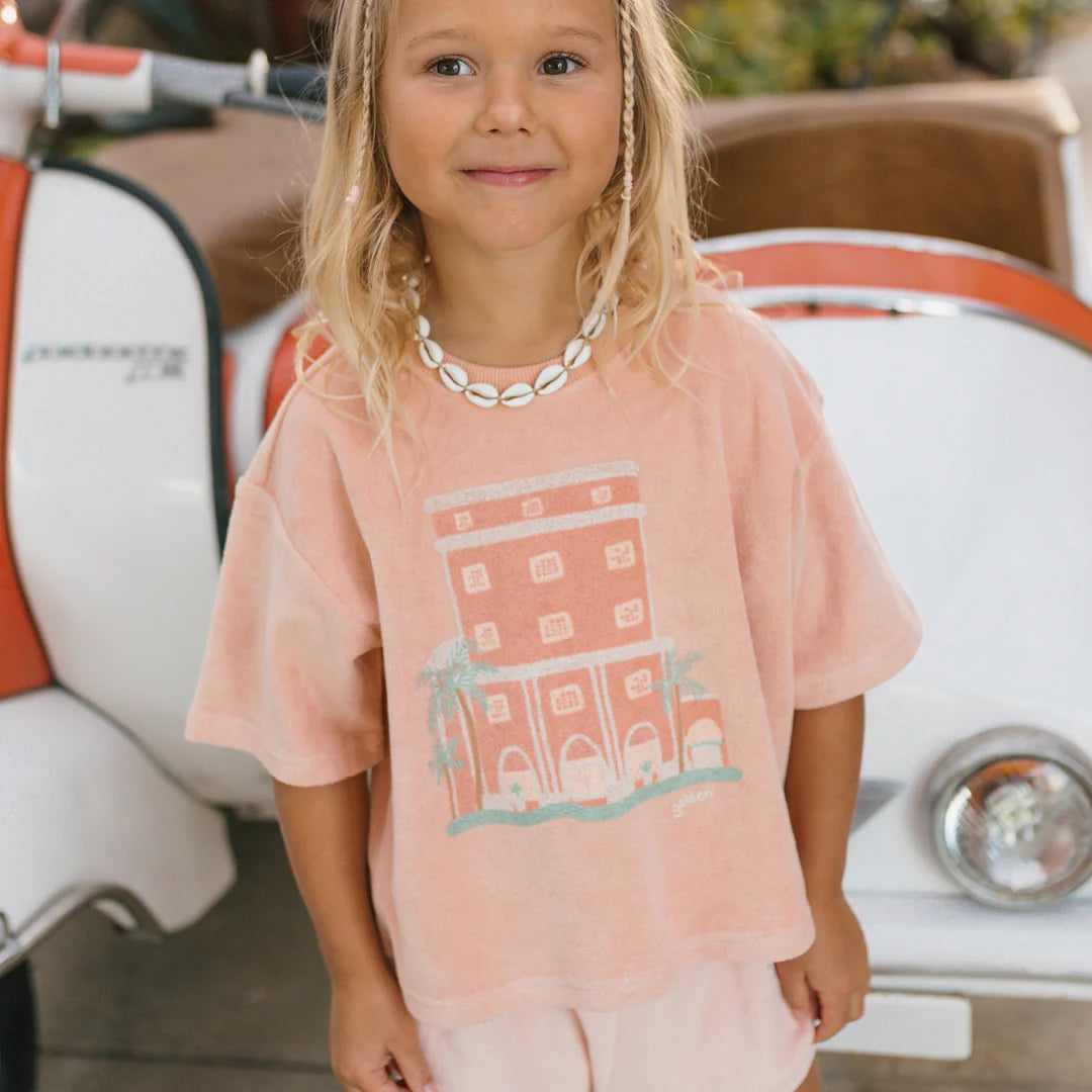 A young child with long hair and a shell necklace smiles at the camera, wearing a peach-colored Room Service Mid Sleeve Tee Terry Towel Spanish Villa from GOLDEN CHILDREN, featuring a graphic of a building. In the background, there's a white and orange vehicle, reminiscent of the ones you'd see parked in front of the Colony Hotel.