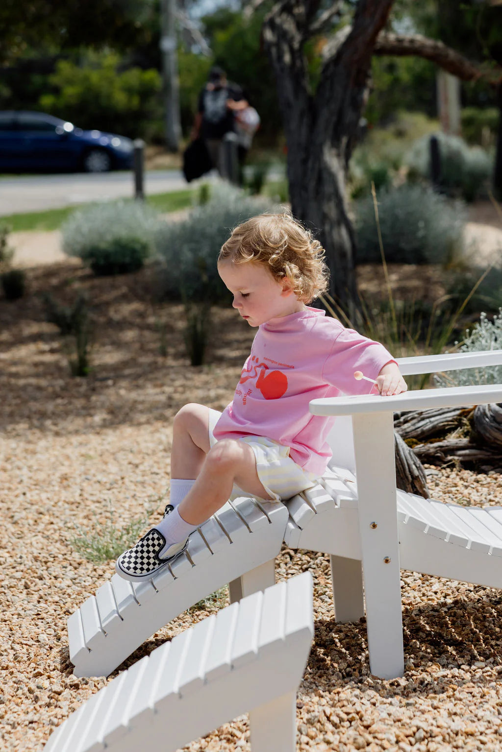 A young child with curly hair sits on white playground equipment, wearing an oversized fit pink Il Sole Tee by SUNDAY SIBLINGS with a red heart, white shorts, and checkered shoes. The scene features trees, plants, and a parked car in the background.