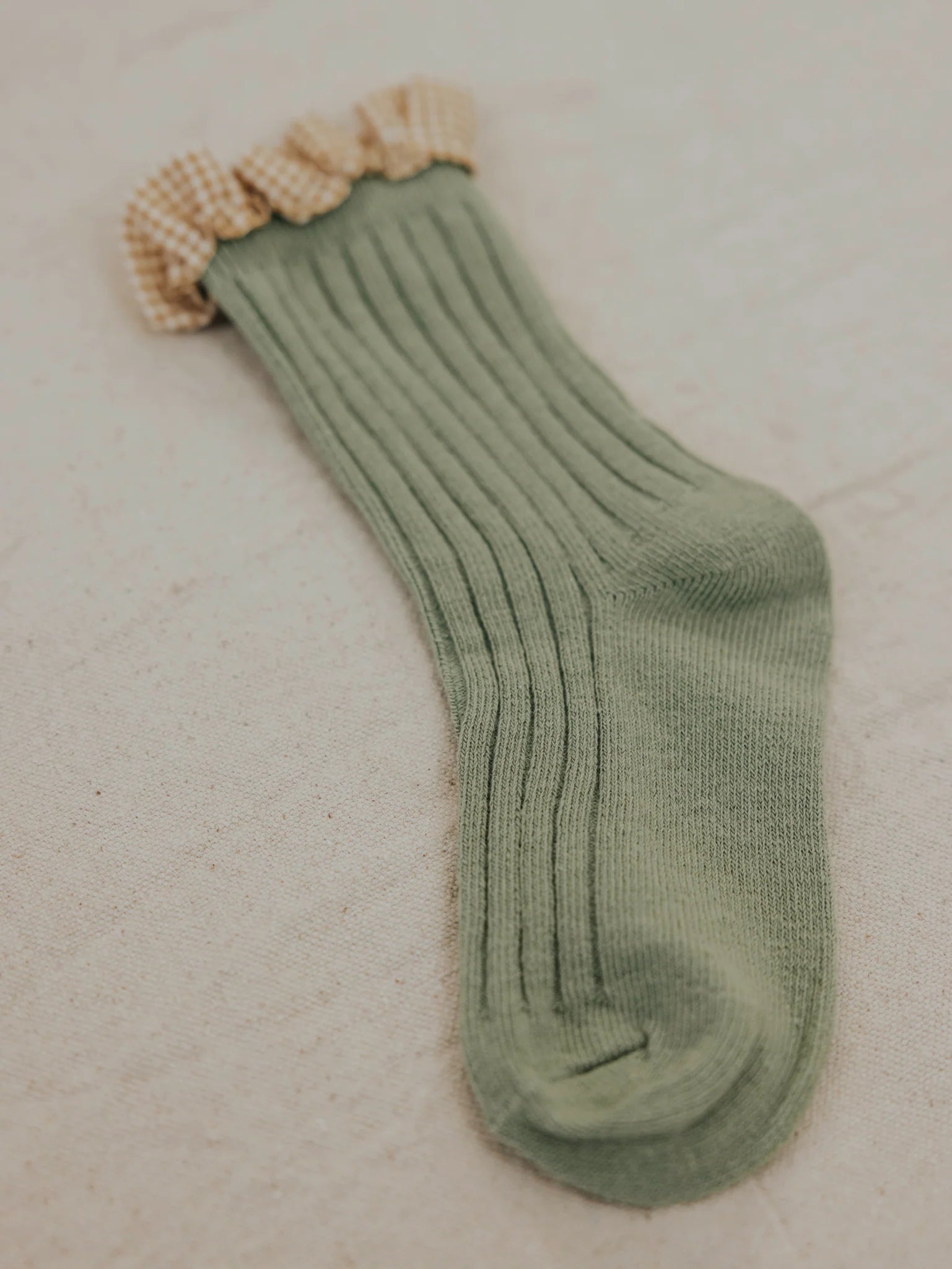 Single Frilly Socks Avocado by VALENCIA BYRON BAY, featuring a green ribbed cotton design with a beige and white gingham frill at the top, laid flat on a light-colored surface. Perfect for those who love frilly socks with a touch of charm.