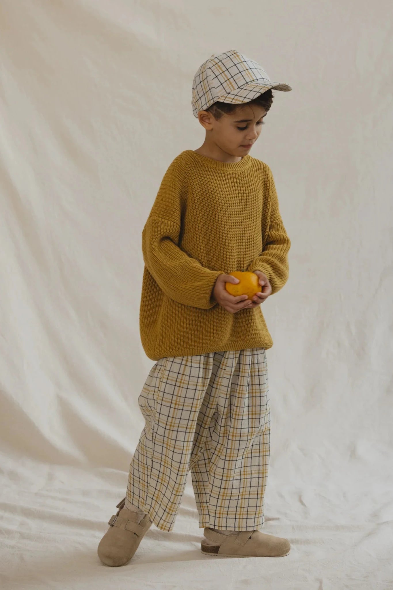 A child in a mustard yellow sweater, VALENCIA BYRON BAY's unisex Rio Pant Picnic in plaid with an elastic waistband (Size 6), and a matching plaid cap holds an orange fruit while standing on a light-colored fabric backdrop.