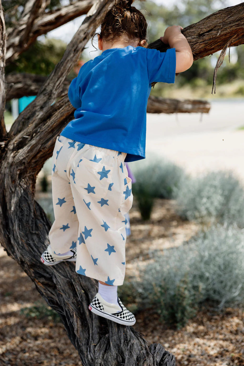 A child in a blue shirt and SUNDAY SIBLINGS' Dusty Star Pants, featuring an elastic waistband, climbs a tree branch. The checkered sneakers add flair to their adventurous spirit against a serene backdrop of greenery and a blurred pathway.