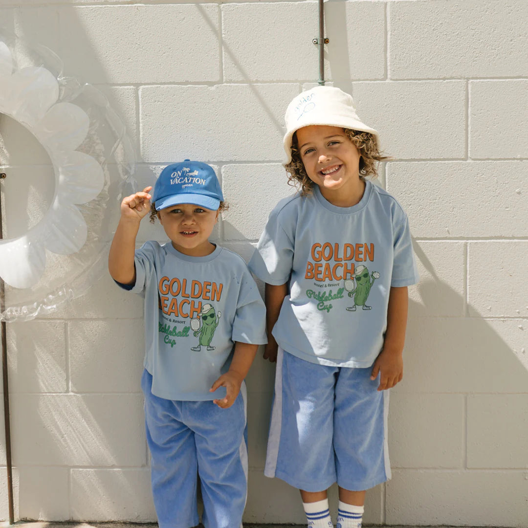 In front of a white wall, two children smile while wearing relaxed fit "Golden Beach Mid Sleeve Tee Palm Blue" shirts from the GOLDEN CHILDREN brand. One child wears a blue cap, while the other sports a white bucket hat. A life preserver is nearby, creating the perfect backdrop for sunny pickleball days by the shore.