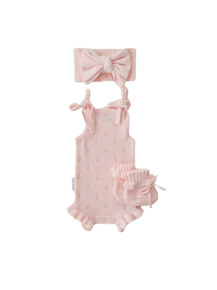 The Frill Bodysuit Primrose by ZIGGY LOU is a baby girl's pink knit romper set adorned with small floral patterns. Made from 100% cotton, the set includes a sleeveless knitted frill bodysuit featuring shoulder tie straps, ruffled leg openings, and a matching headband with a large bow, along with ruffled bloomers.