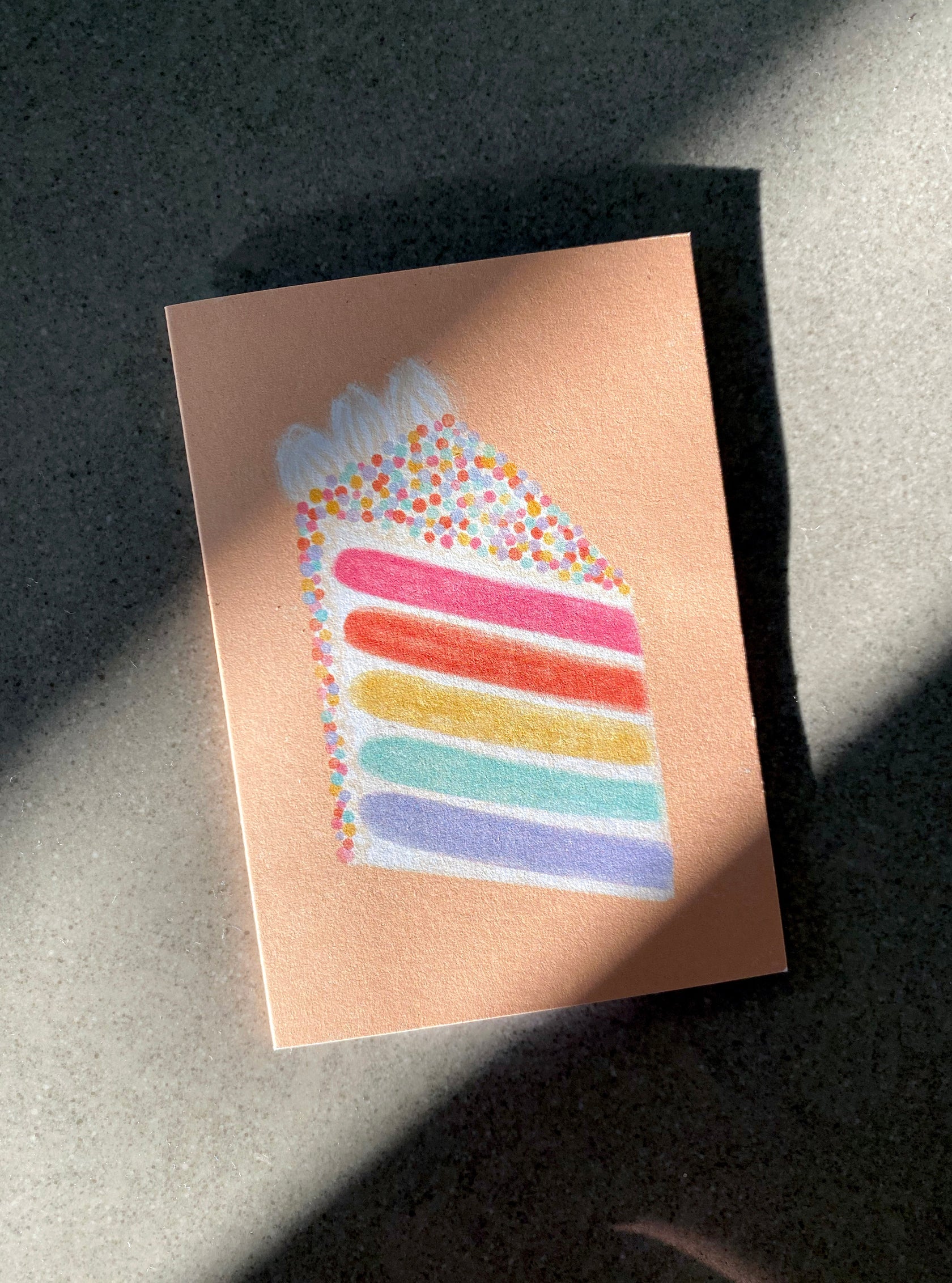 The Rainbow Cake Mini Card by LAUREN SISSONS STUDIO features an illustration of a rainbow-layered cake slice with white frosting and colorful sprinkles. Crafted from recycled paper, the card is set against a gray surface with streaks of sunlight casting shadows over it.