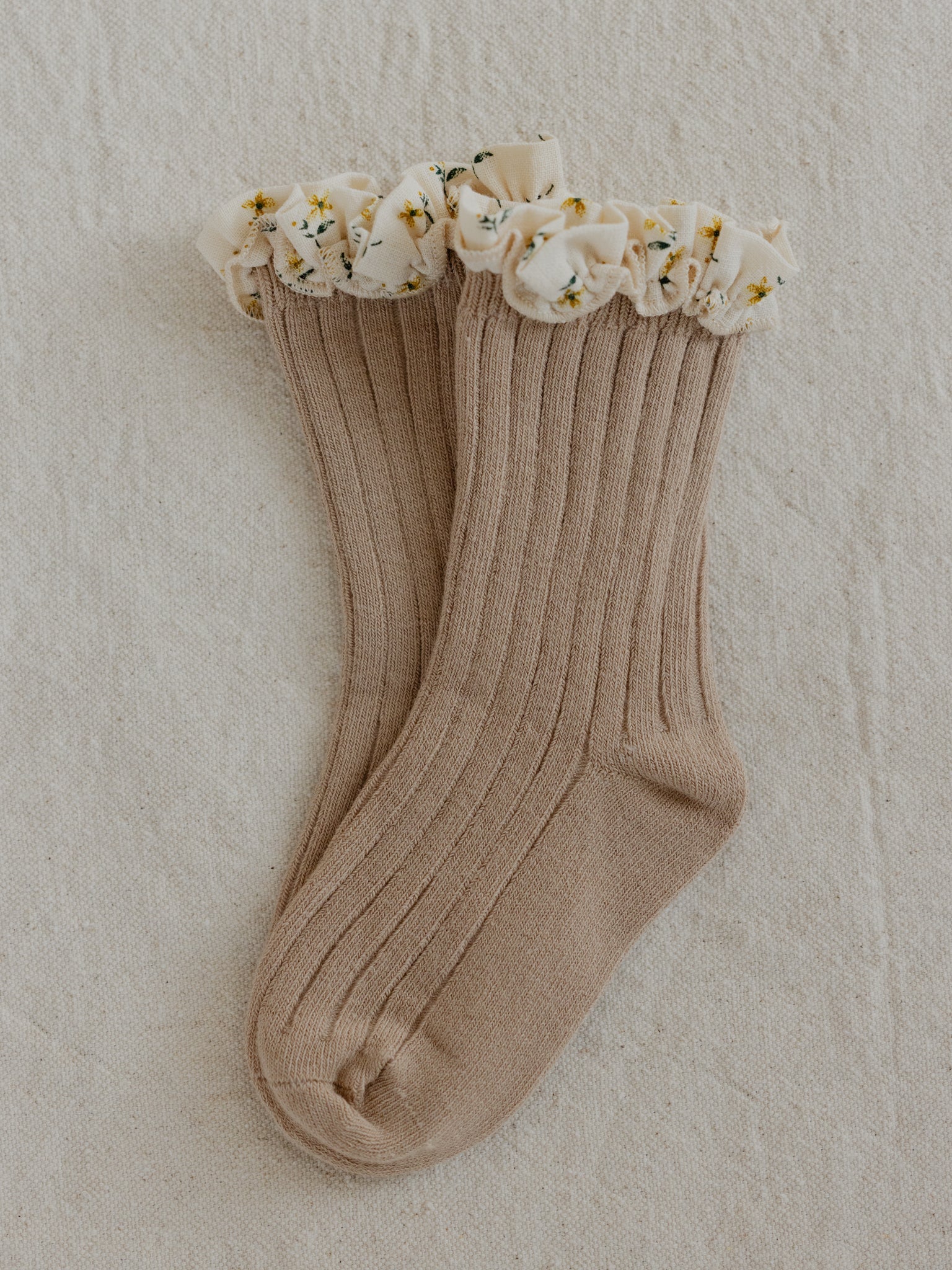 A pair of beige ribbed Marigold Socks Clay by VALENCIA BYRON BAY, featuring printed frill tops with small white flowers and green leaves adorning the ruffles, are laid flat on a textured, neutral-colored surface, capturing the serene essence of Byron Bay.