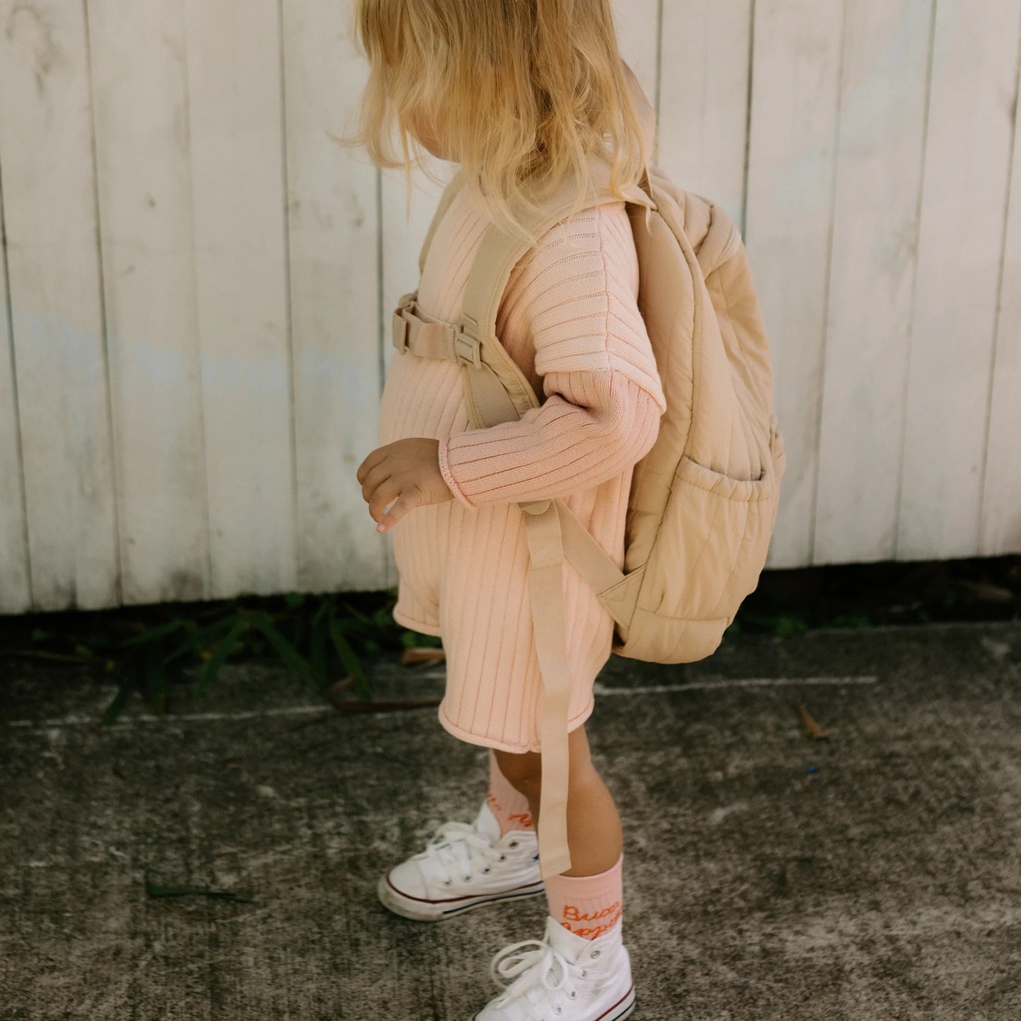 A young child with blonde hair stands facing away from the camera, wearing the Golden Knit Romper Strawberry Sundae by GOLDEN CHILDREN. The light pink ribbed knit romper, made of 100% cotton, is paired with a beige backpack and white sneakers. They are standing on a concrete surface in front of a wooden fence.