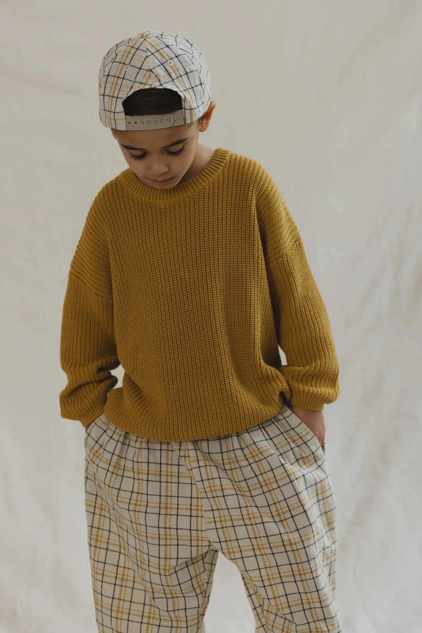 A young child gazes downward, hands in pockets, dressed in VALENCIA BYRON BAY's unisex Rio Pant Picnic - Size 6. Their mustard sweater and matching plaid pants with an elasticated waistband are complemented by a cap, all set against a simple backdrop.