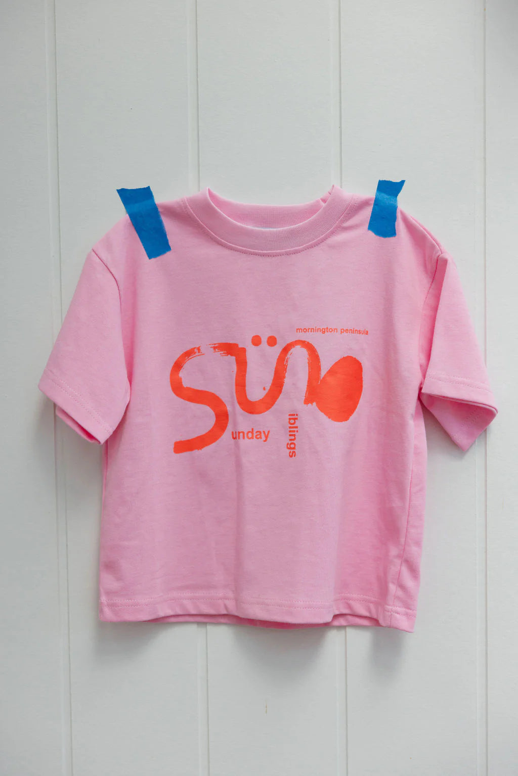 Introducing the Sunday Siblings Il Sole Tee: a pink/red children's T-shirt made from 100% cotton, featuring "Sun" in bold orange and "sunday things" in smaller print. Its oversized fit adds a trendy touch as it hangs on a white wall with blue tape.